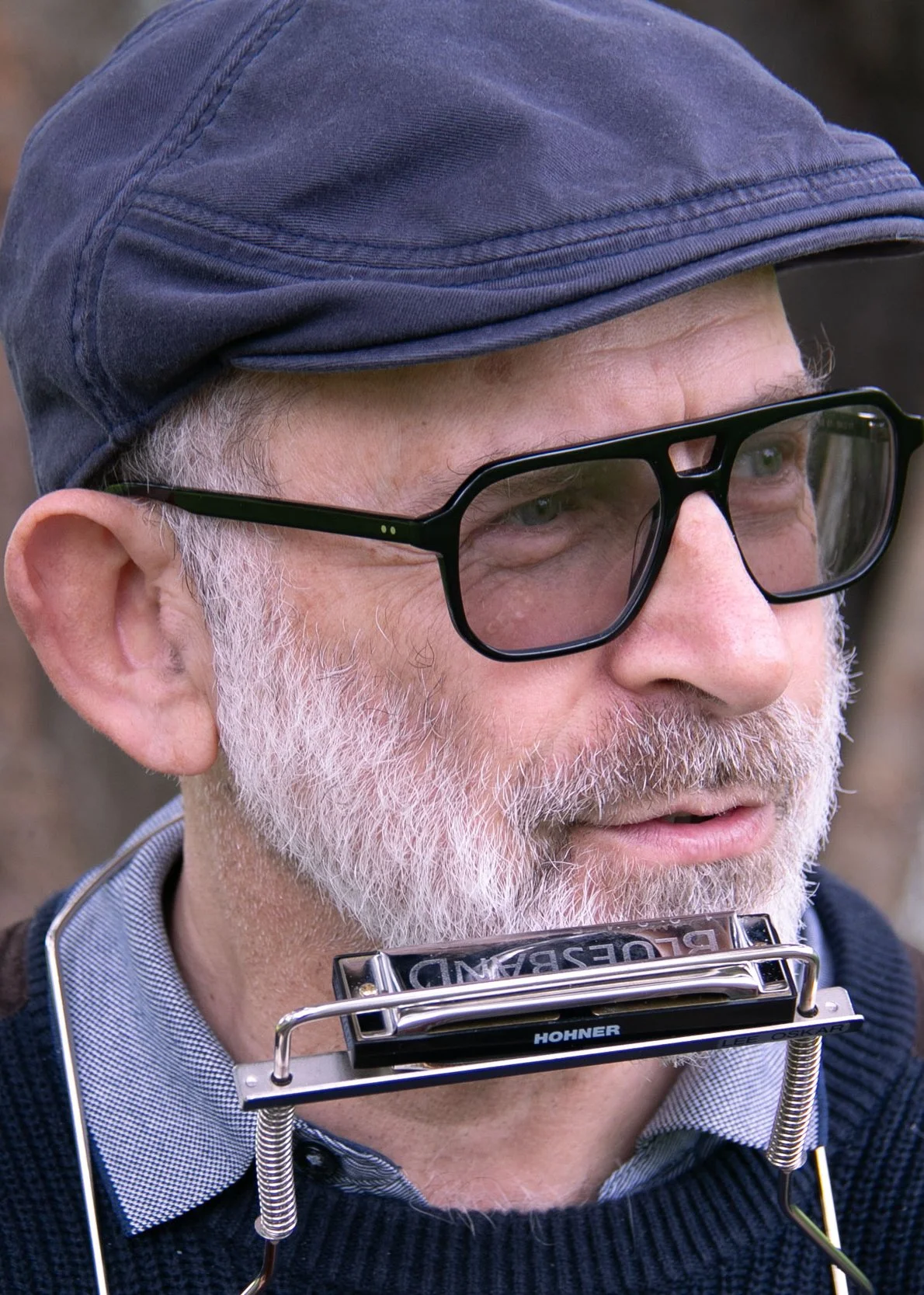 Close-up of an older man with a white beard wearing glasses, a hat, and a harmonica holder around his neck.