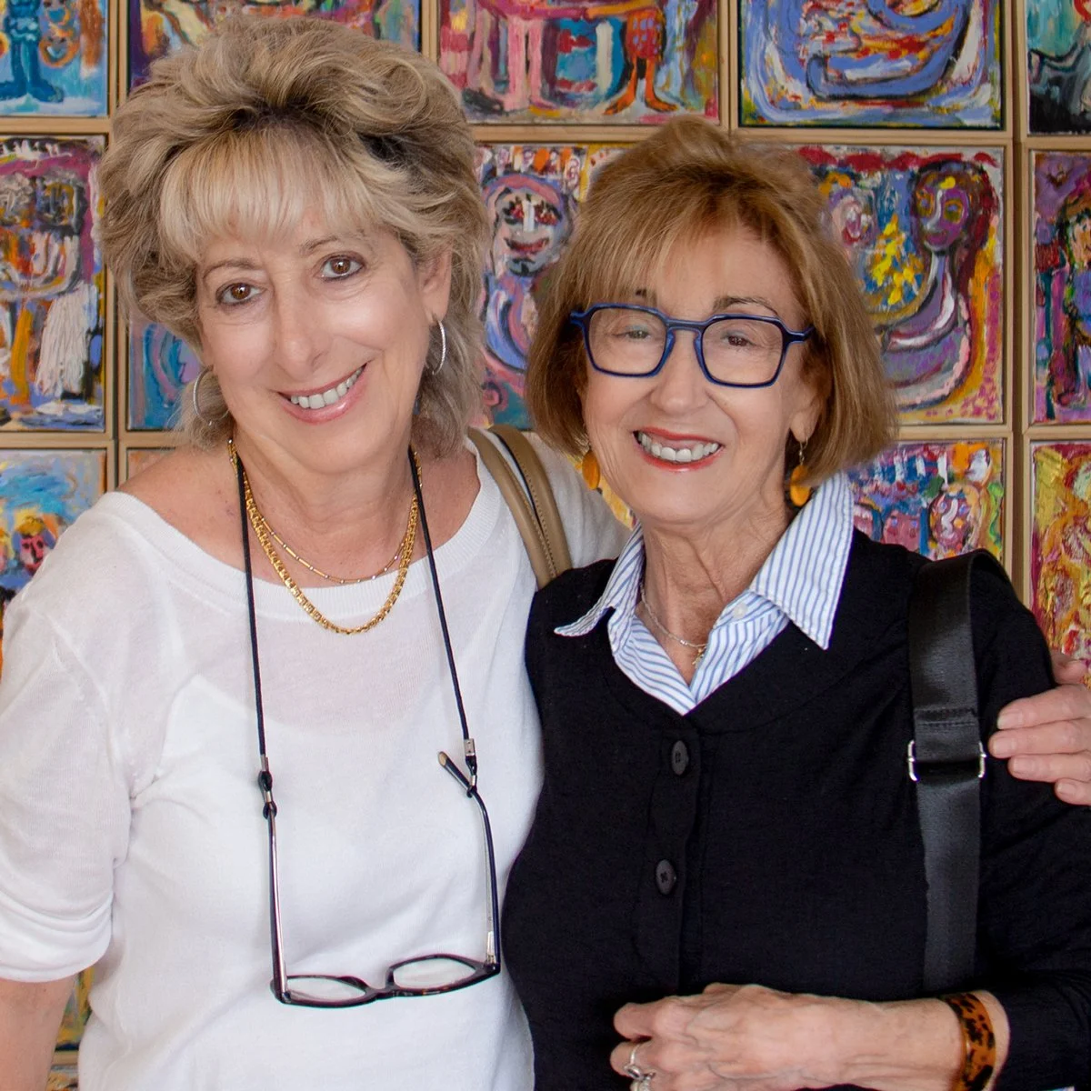Two women smiling and standing close together in front of colorful abstract artwork.