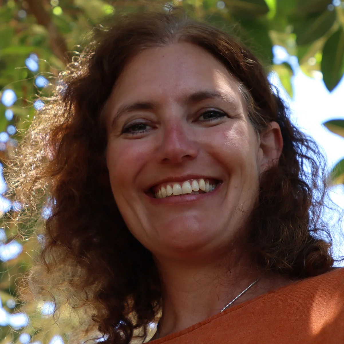Close-up of a smiling woman with curly brown hair, outdoors with sunlight and green leaves in the background.