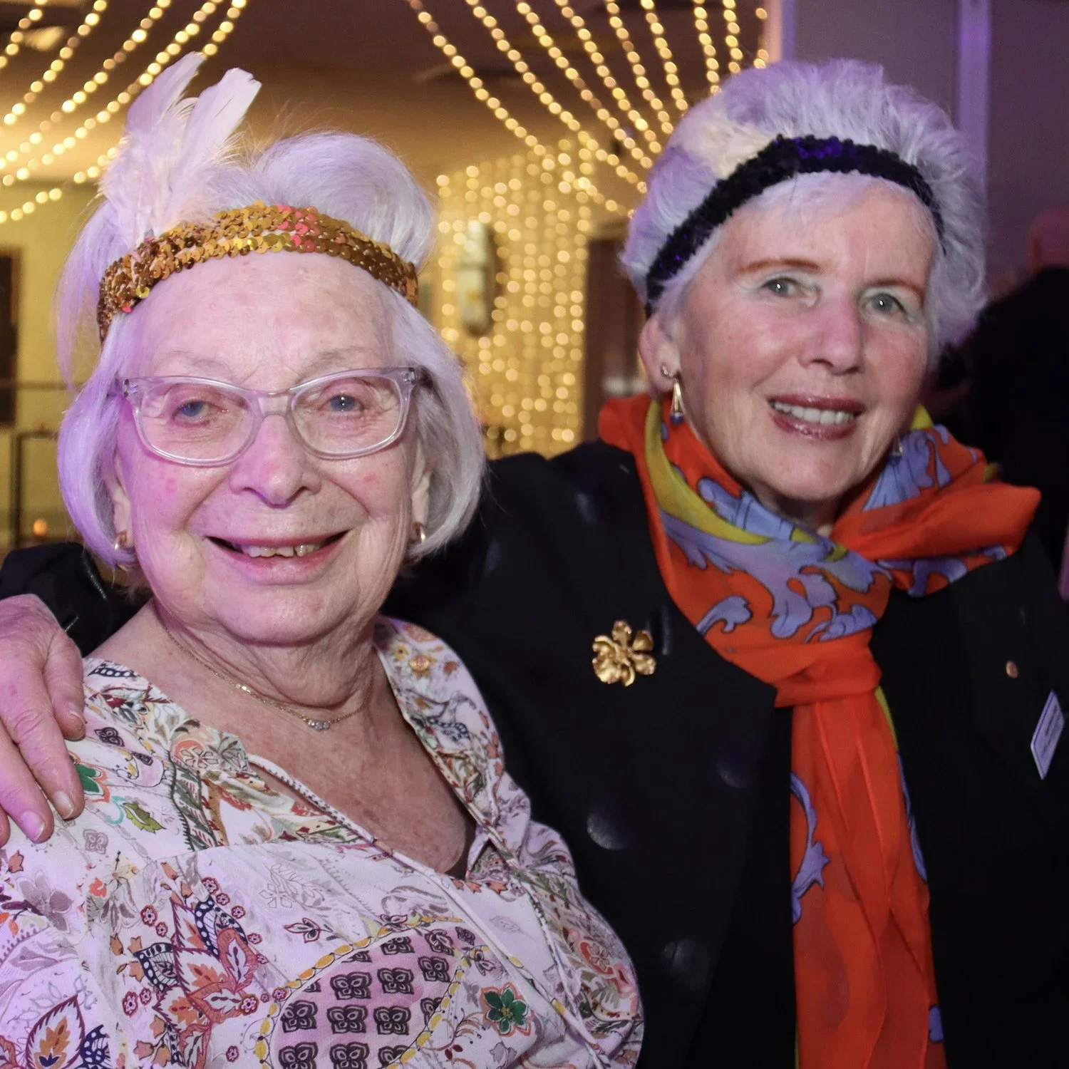 Two elderly women posing together at a festive event with string lights in the background. They are smiling and dressed colorfully, one with glasses and a feathered headband, the other wearing a vibrant scarf and a flower-shaped brooch.