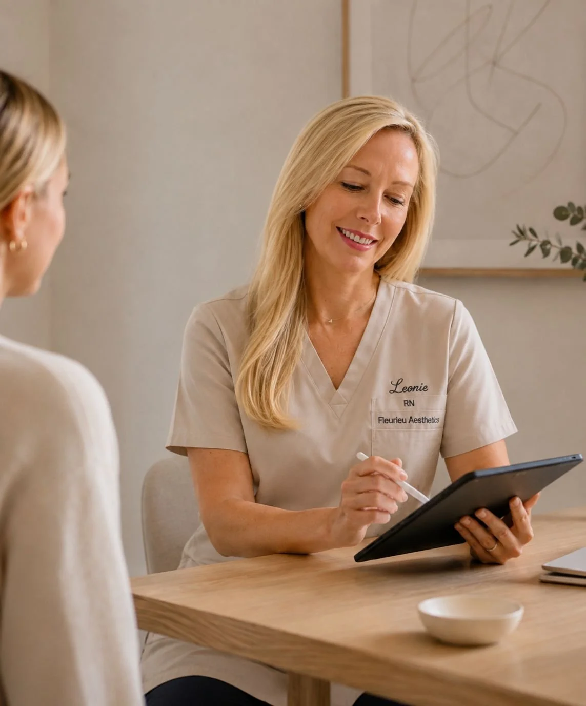 A woman with blonde hair wearing a beige dental or aesthetic professional uniform, smiling and looking at a tablet, sitting at a wooden table in a relaxing office or consultation room.