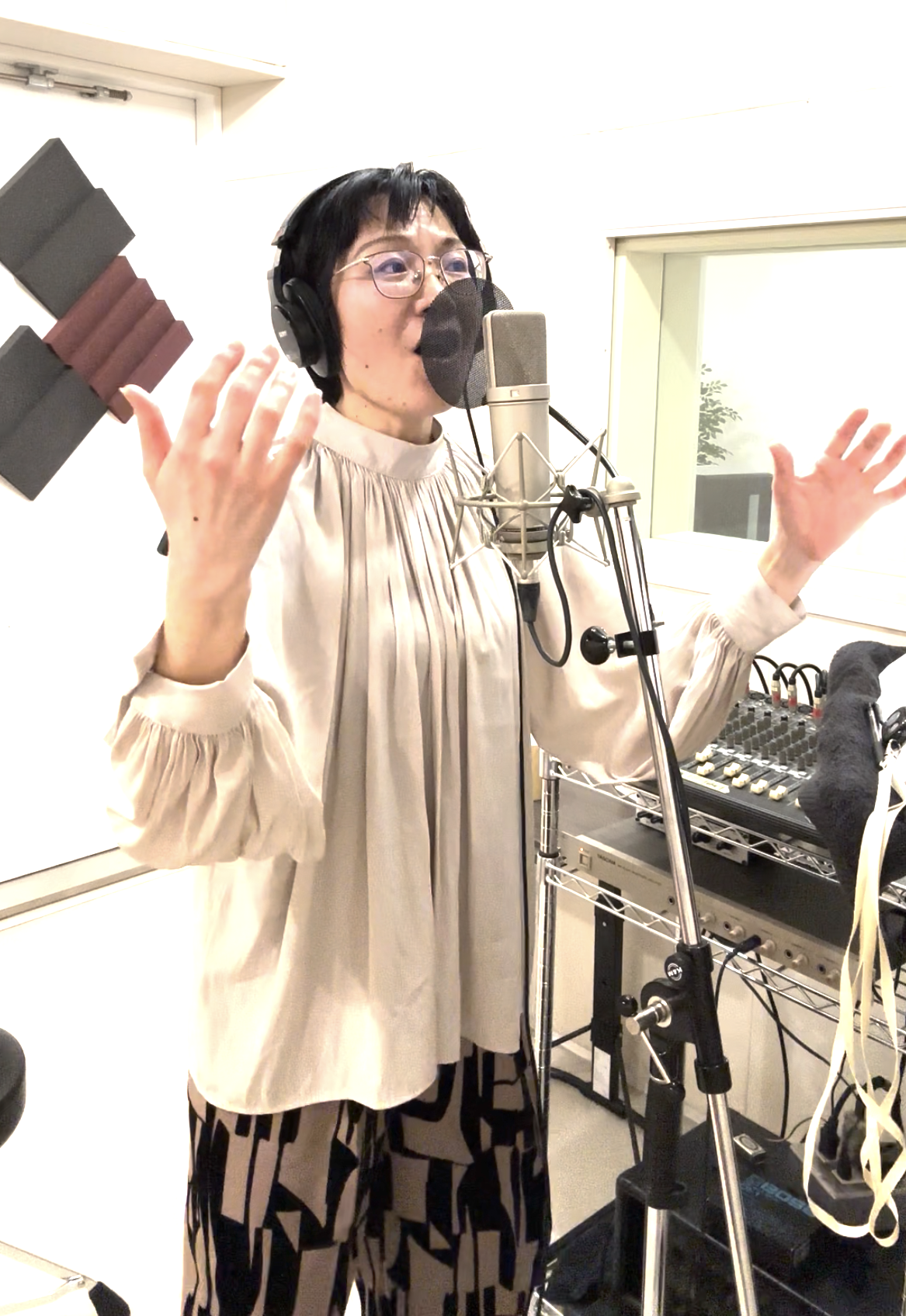 スタジオでマイクに向かって歌う女性。ヘッドフォンをつけ、音響機器の前に立っている。