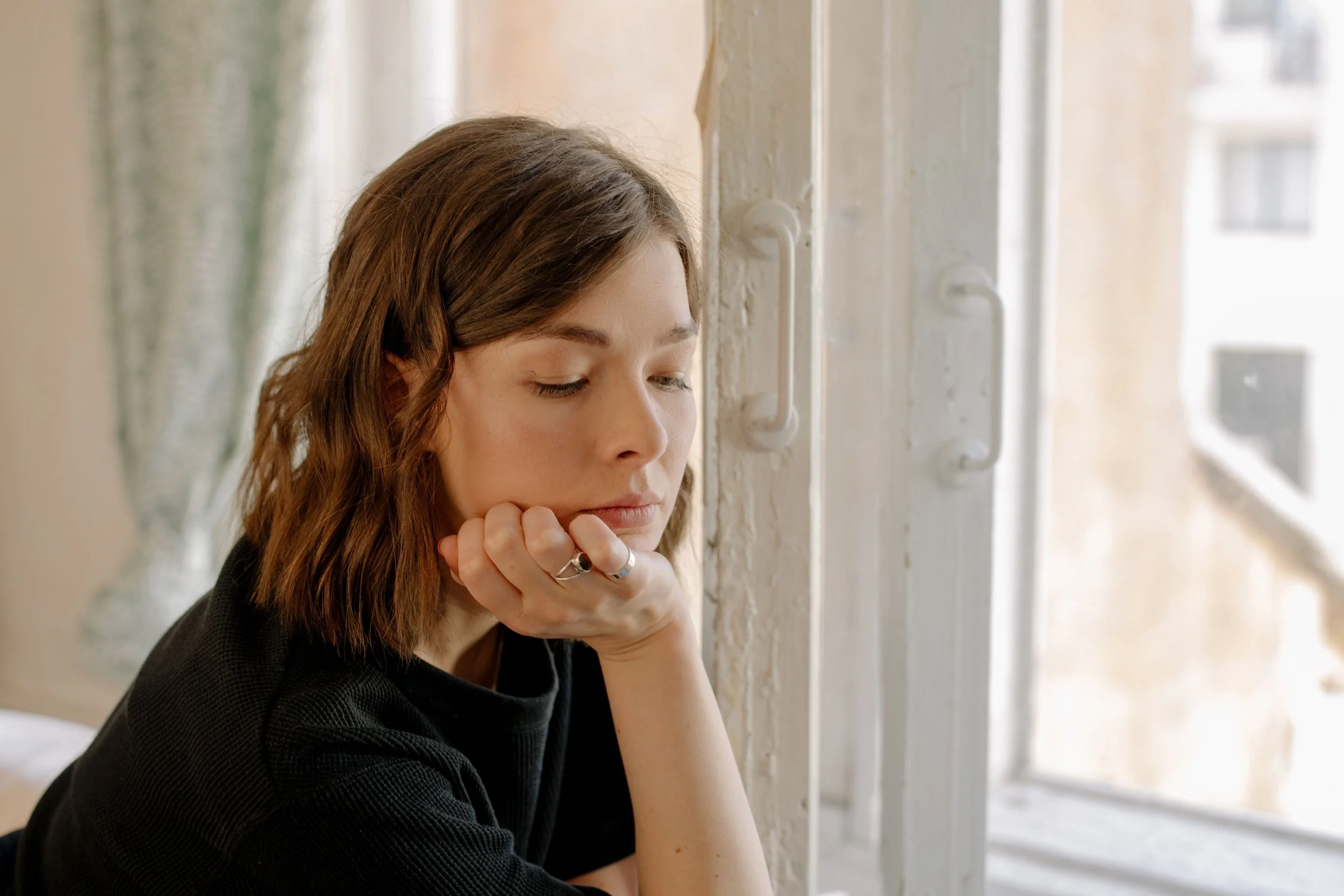 A young woman with wavy brown hair sitting near a window, with her chin resting on her hand, appearing thoughtful or contemplative.