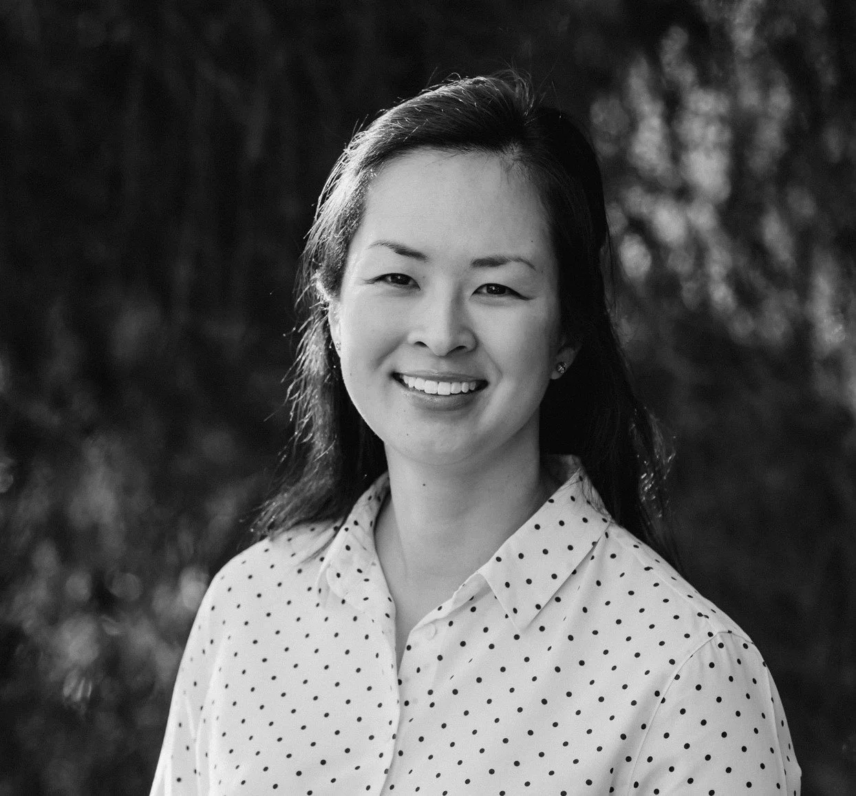Black and white portrait of a young Asian woman with shoulder-length hair, smiling, wearing a polka dot blouse, outdoors with blurred trees in the background.