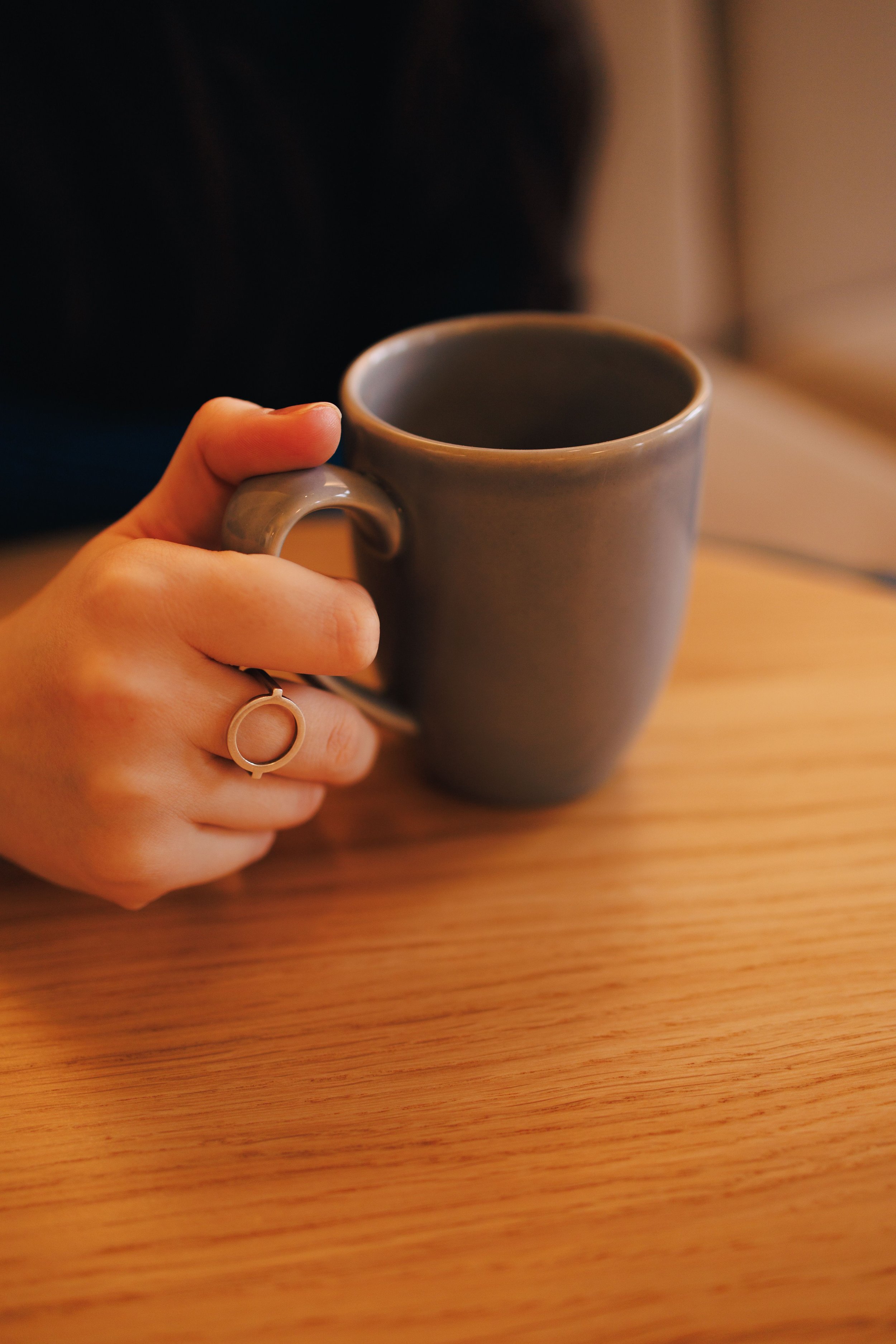 A person's hand holding a grey ceramic mug with a plain wooden table in the background.