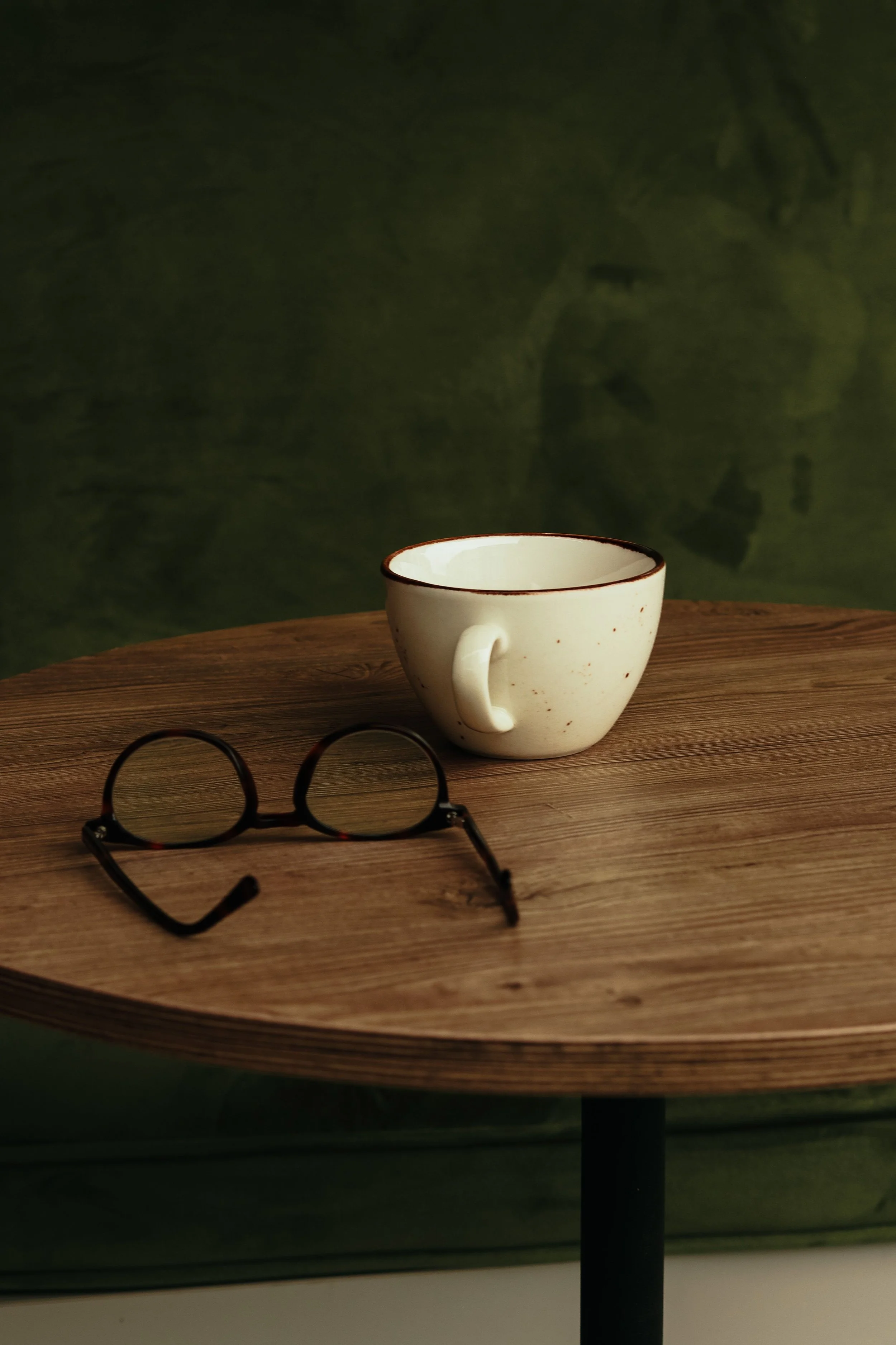 A white ceramic cup with a dark rim sits on a round wooden table. A pair of black eyeglasses with tortoiseshell temples is in the foreground on the table. The background is a dark green wall.