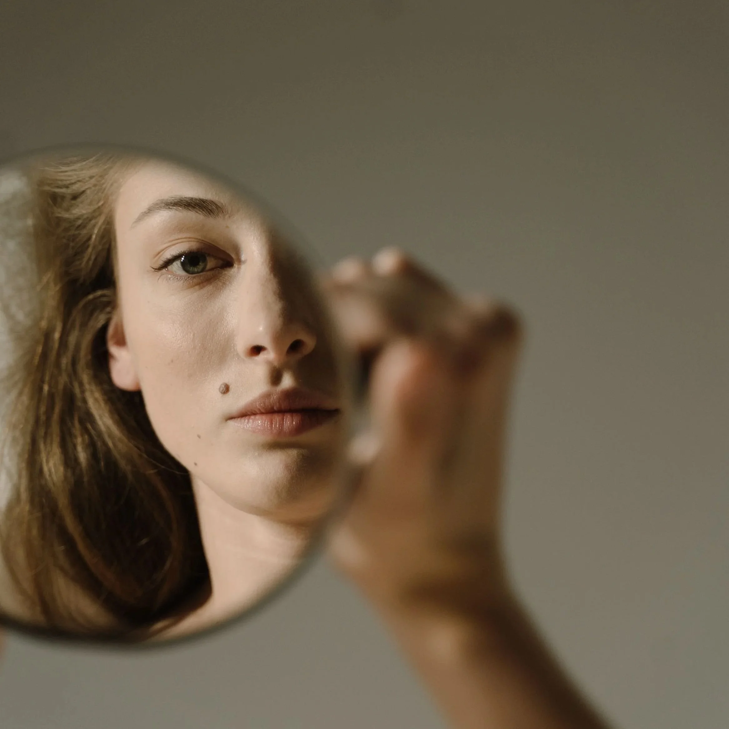 Close-up of a woman with fair skin, brown hair, and a beauty spot on her face, posing in front of a mirror