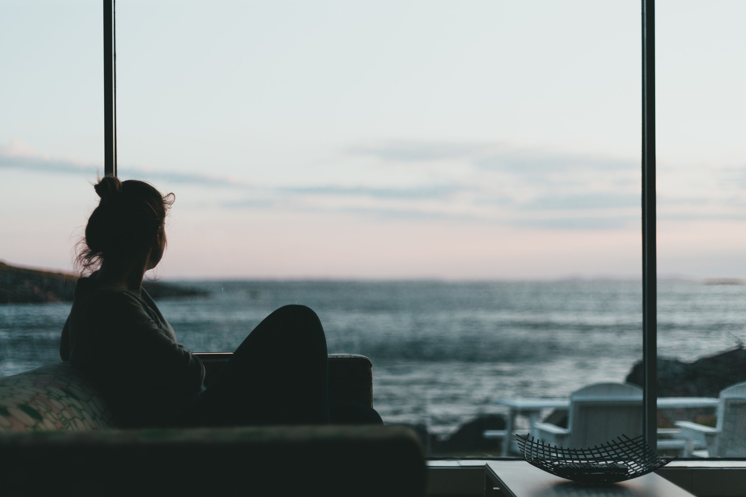 Person with hair in a bun sitting indoors near large windows, with a view of the ocean and sky in the background.