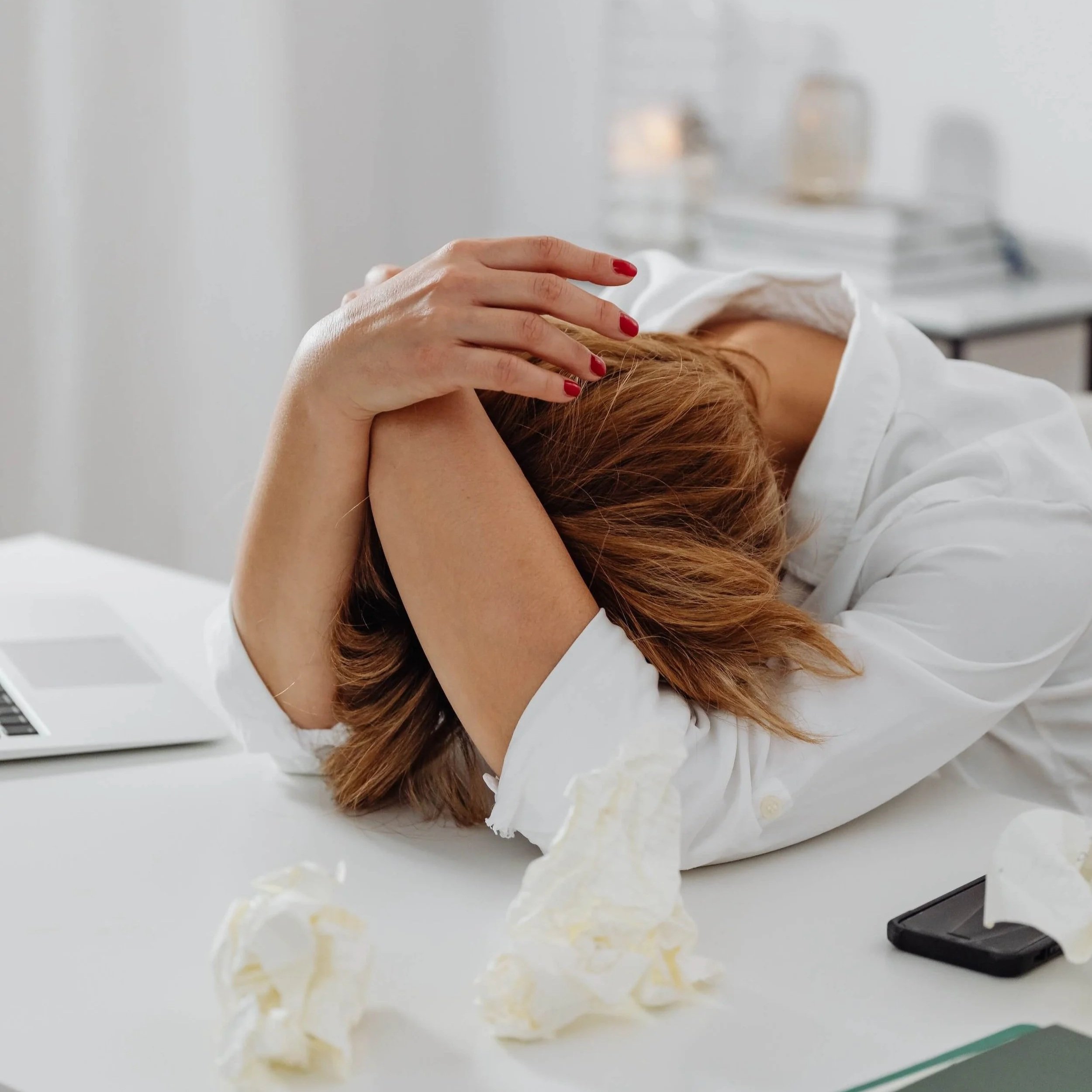 A person with red hair and red nail polish resting their head on their arm on a white table. There is a piece of cake or pastry with yellow filling and white frosting on the table in front of them.