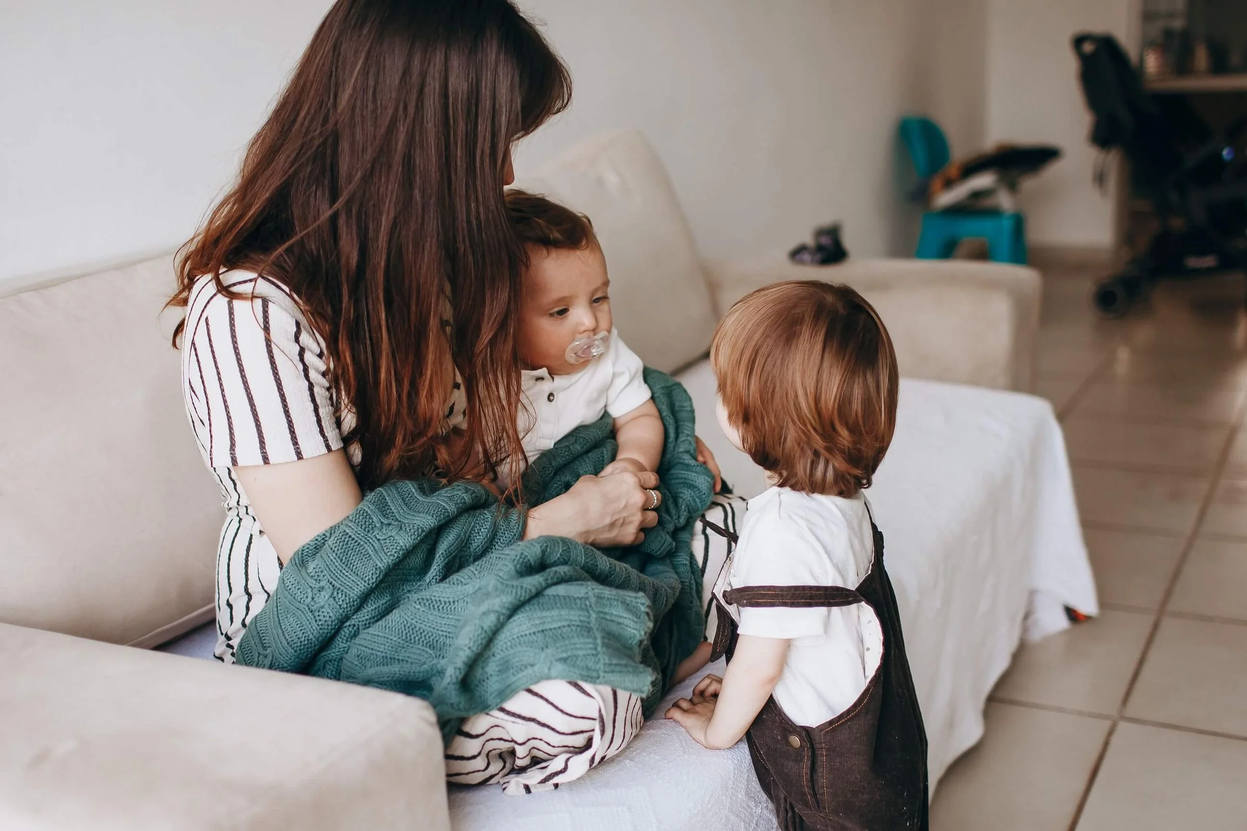 A woman with long brown hair sitting on a sofa holding a young child with a pacifier in their mouth, while another small child with red hair stands nearby, engaging with the woman and the child.