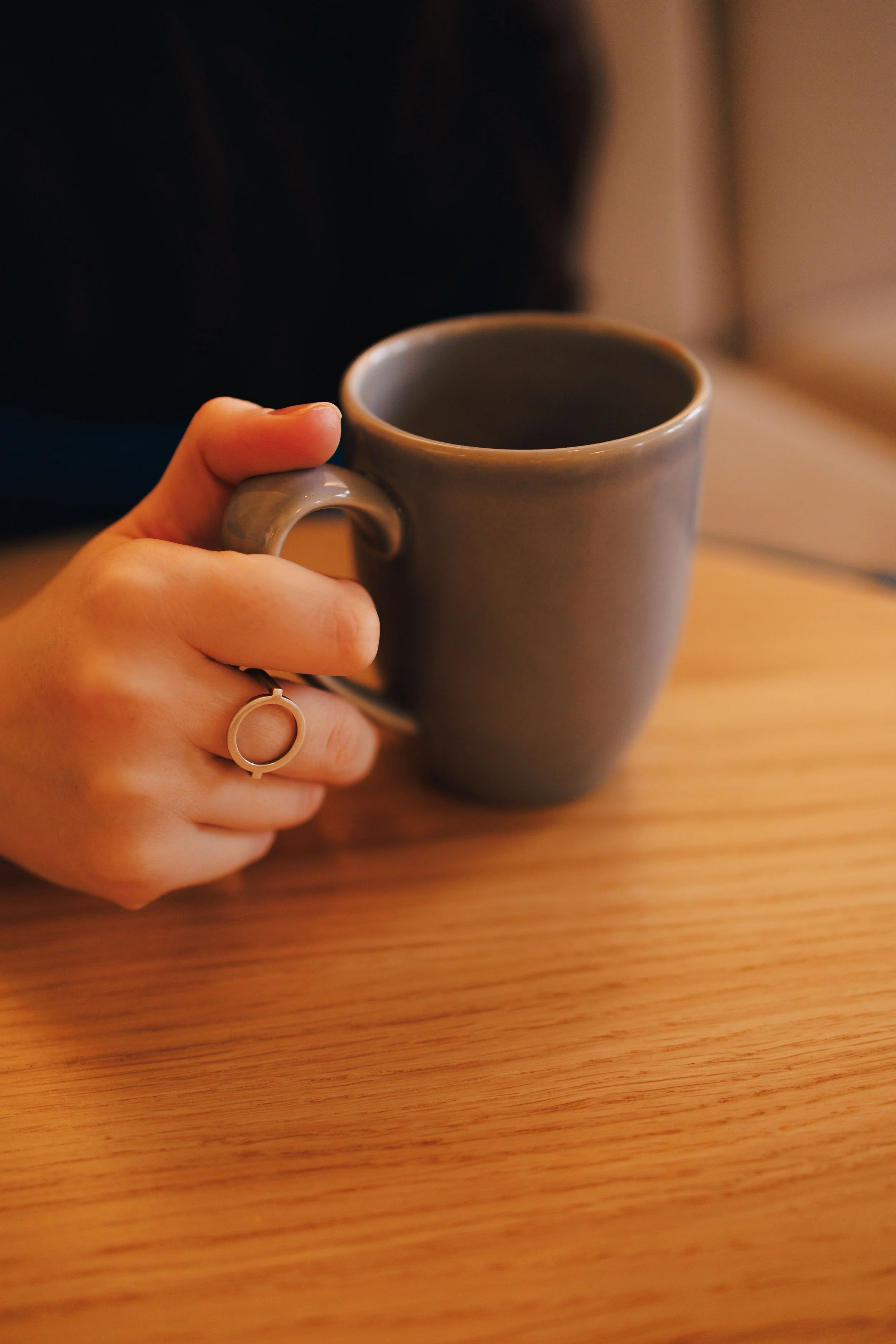 A person holding a gray ceramic coffee mug in their left hand, sitting on a wooden surface with a blurred background.