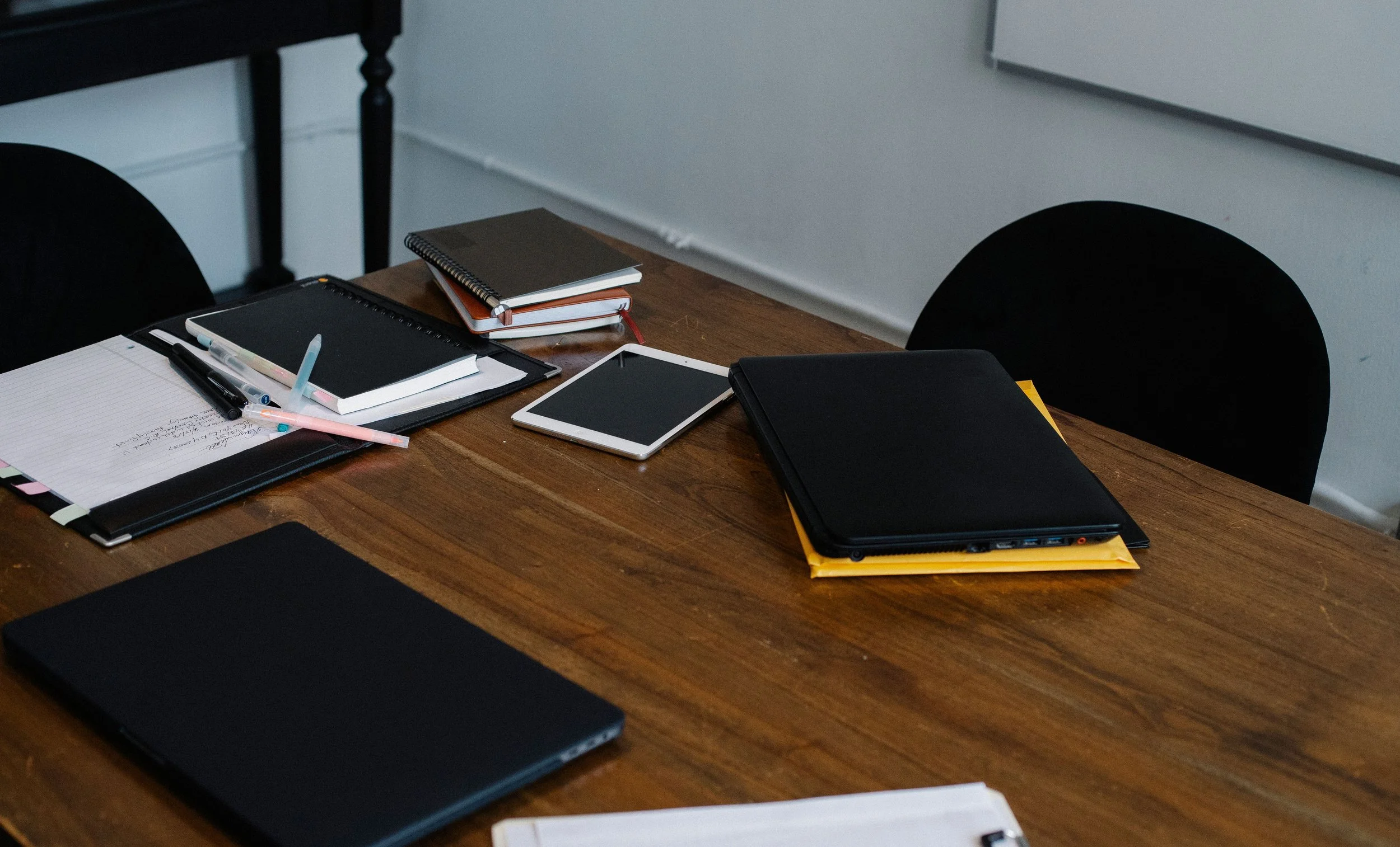 A wooden desk with notebooks, folders, a tablet, a laptop, pens, and papers, in a classroom or office setting.