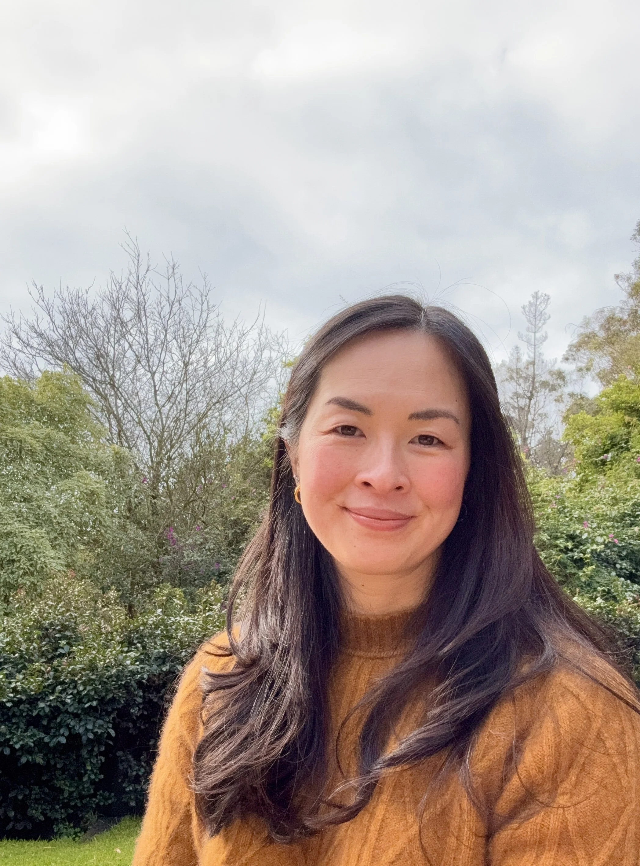 A woman with long dark hair taking a selfie outdoors, with trees and bushes in the background on a cloudy day.