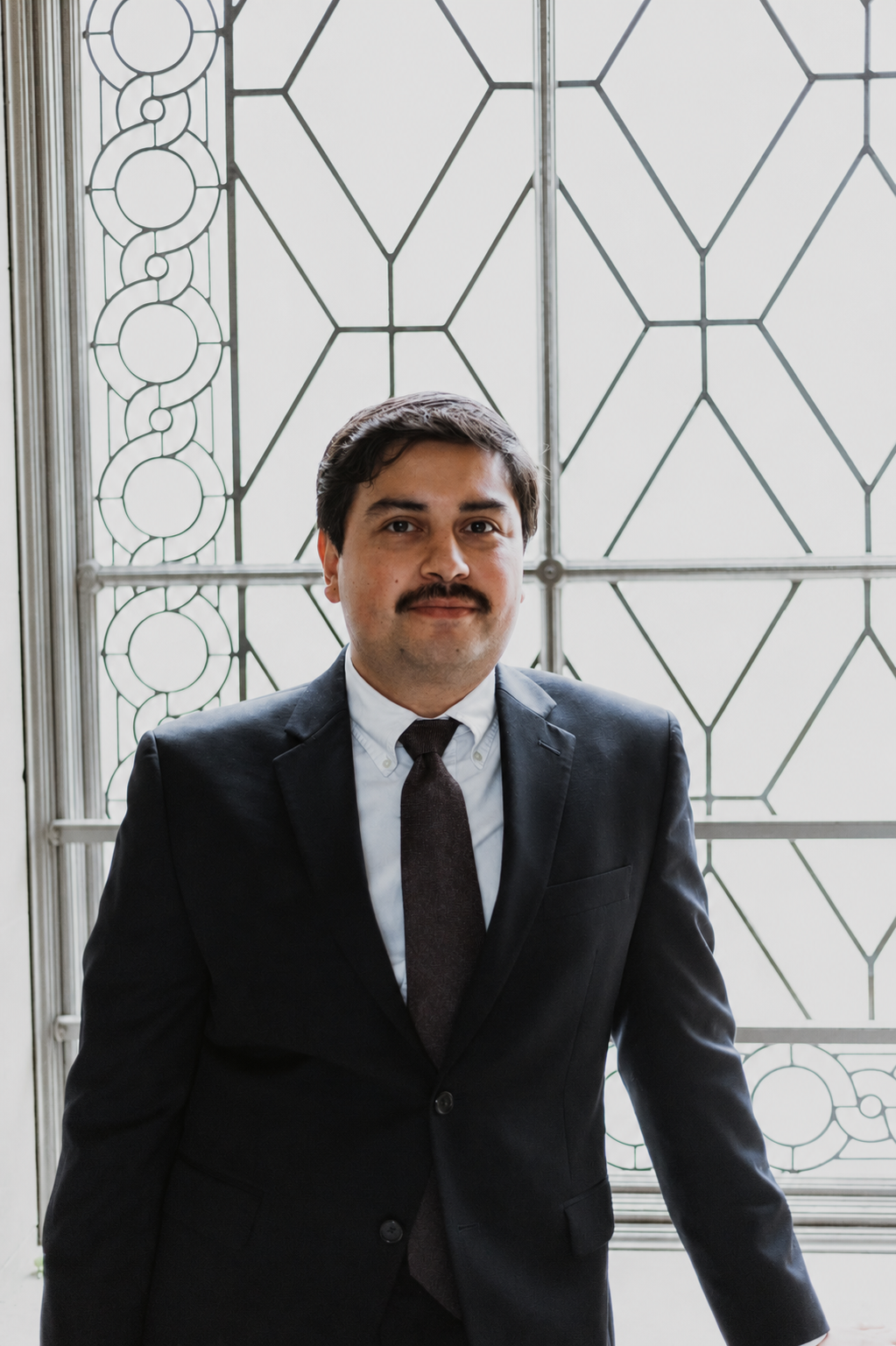 A man with dark hair, a mustache, wearing a dark suit, white shirt, and dark tie, standing in front of a decorative metal window or barrier with geometric patterns.