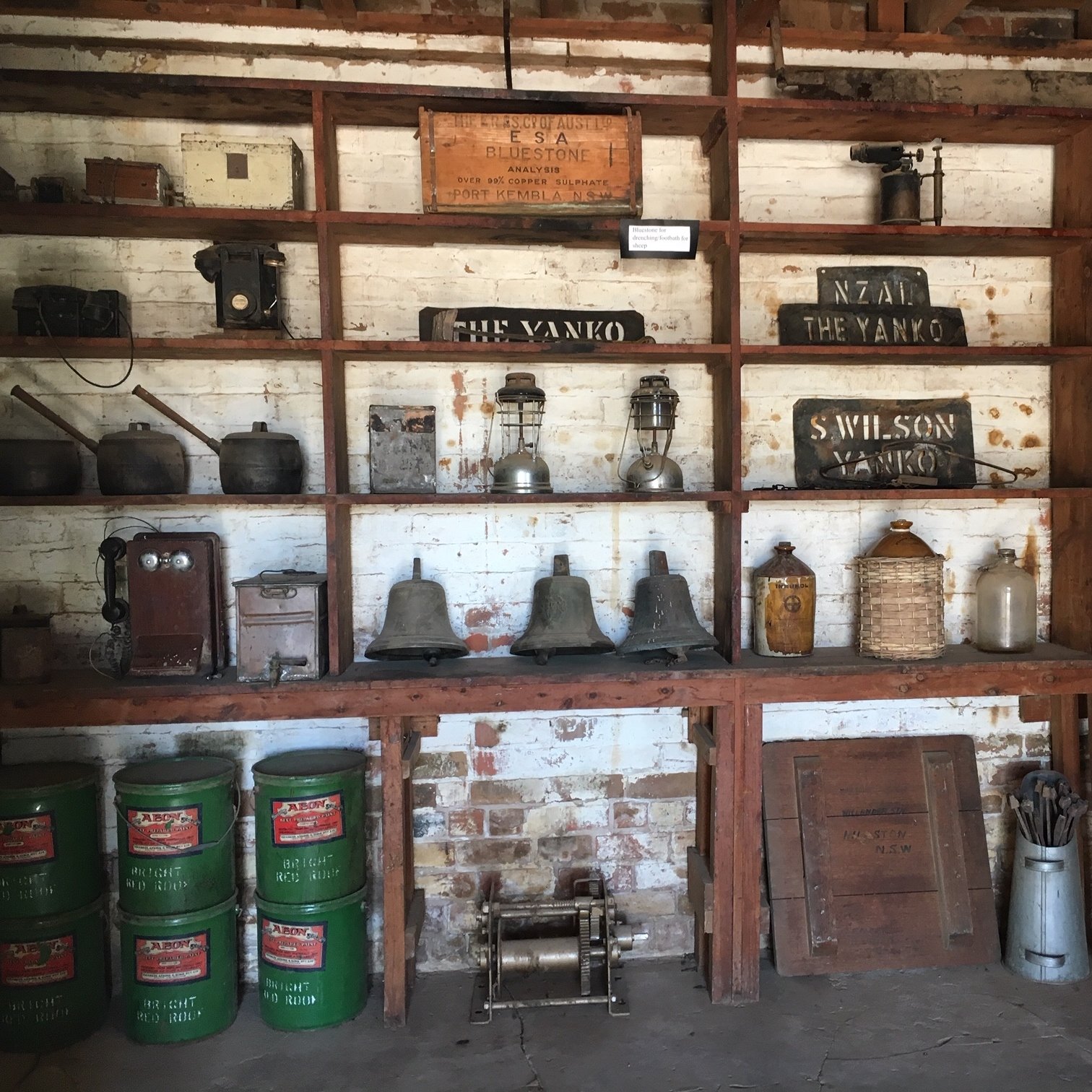 A wooden shelf displaying vintage items including old telephones, lanterns, metal pots, and signs, against a white brick wall in an industrial or museum setting. Inside the historical The Yanko Store