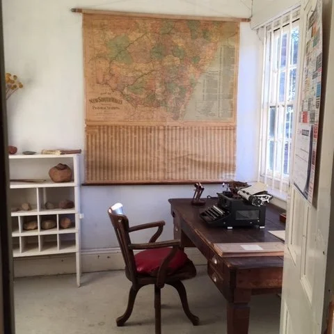 A home office with a vintage map on the wall, a wooden desk with a typewriter, a wooden chair with a red cushion, and a white shelf with stones or decorative objects, next to a window with white blinds.  Inside the historical The Yanko Store