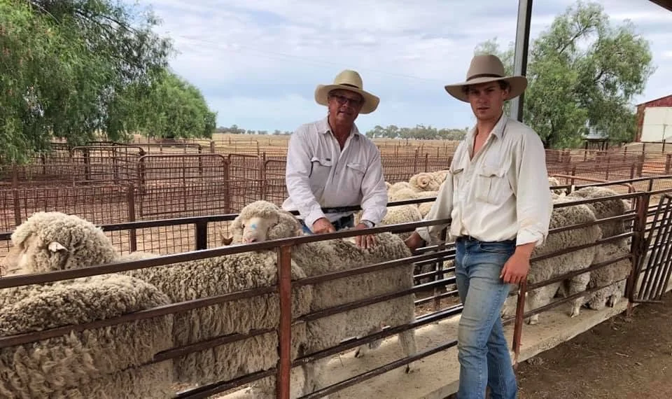 Two men wearing cowboy hats and light-colored shirts are standing in a cattle pen with sheep. One man is older with glasses, and the younger one is leaning on the fence. The background shows a rural landscape with trees and farm buildings.