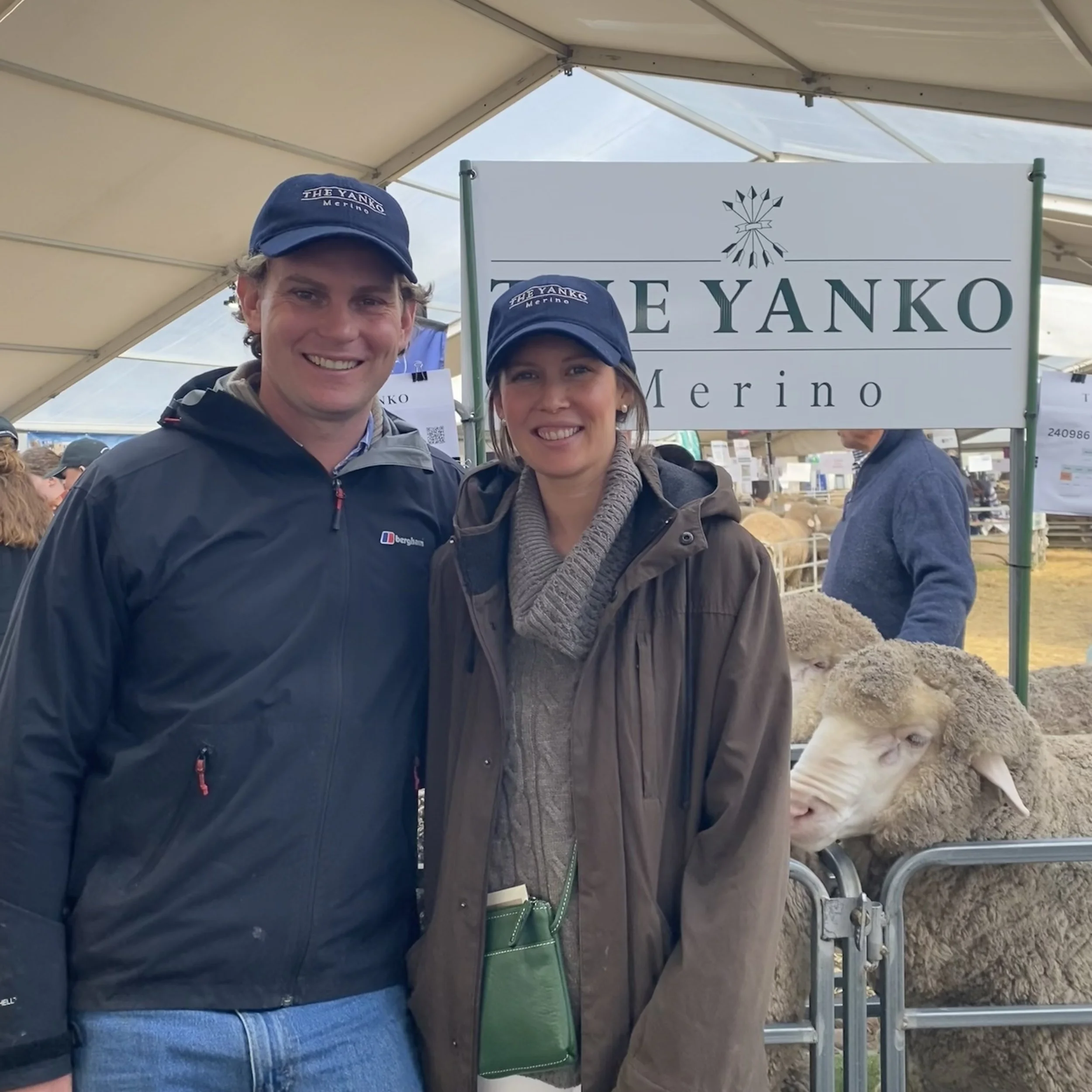 Two smiling people, a man and a woman, standing together under a canopy at an outdoor event. Both are wearing blue hats with the words 'The Yanko Merino' printed on them. The man is wearing a dark jacket and the woman has on a gray scarf and a dark coat. Behind them, there is a sign that reads 'The Yanko Merino' along with a logo. Part of a sheep's head and wool are visible to the right of the image.