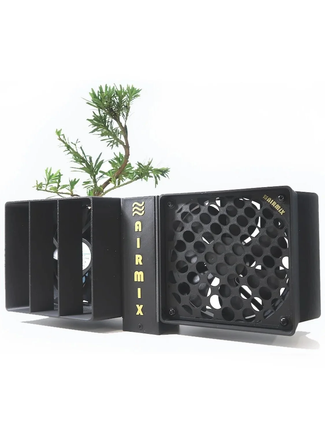 A black 3D-printed AirMix cooling module with yellow lettering, mounted next to a small bonsai plant, set against a plain white background.