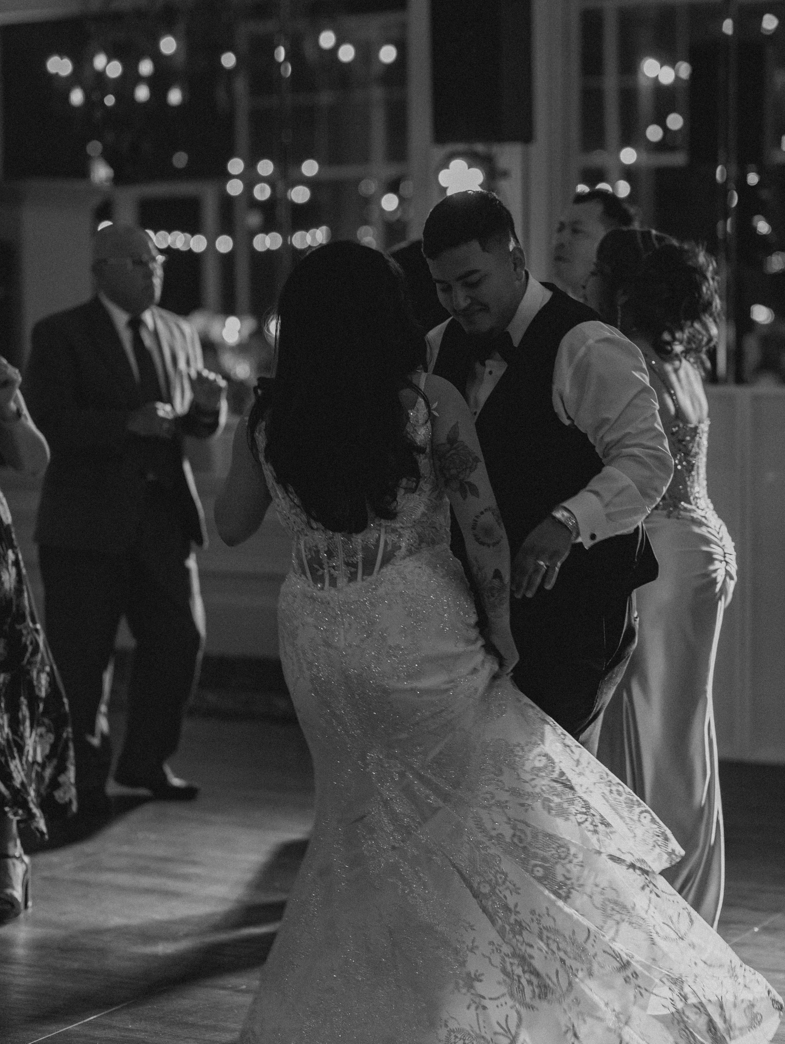 A black and white photo of a wedding reception. A bride and groom are dancing closely, surrounded by guests in formal attire.