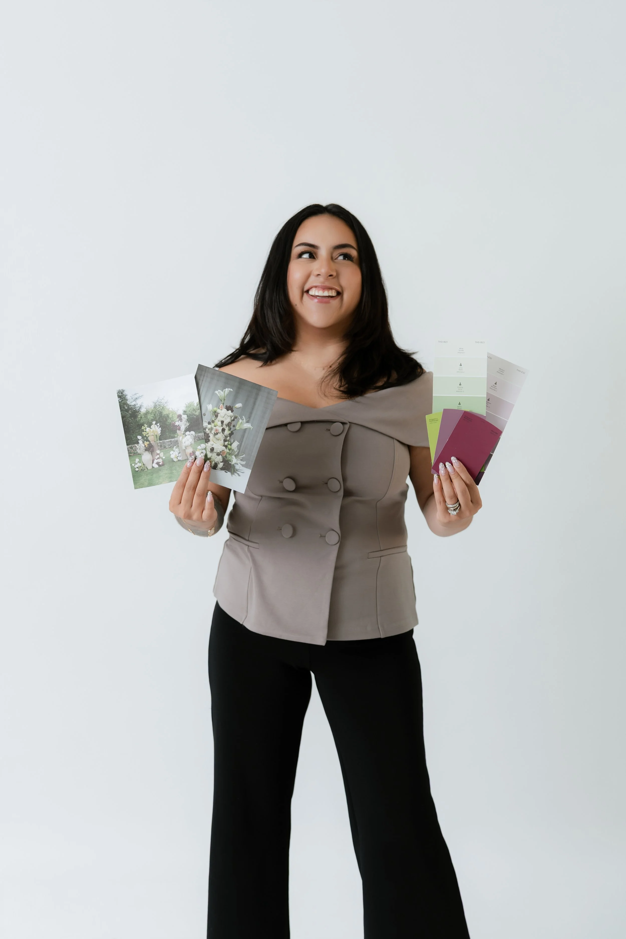 A woman with dark hair smiling and holding color swatches and photographs, standing against a plain white background.