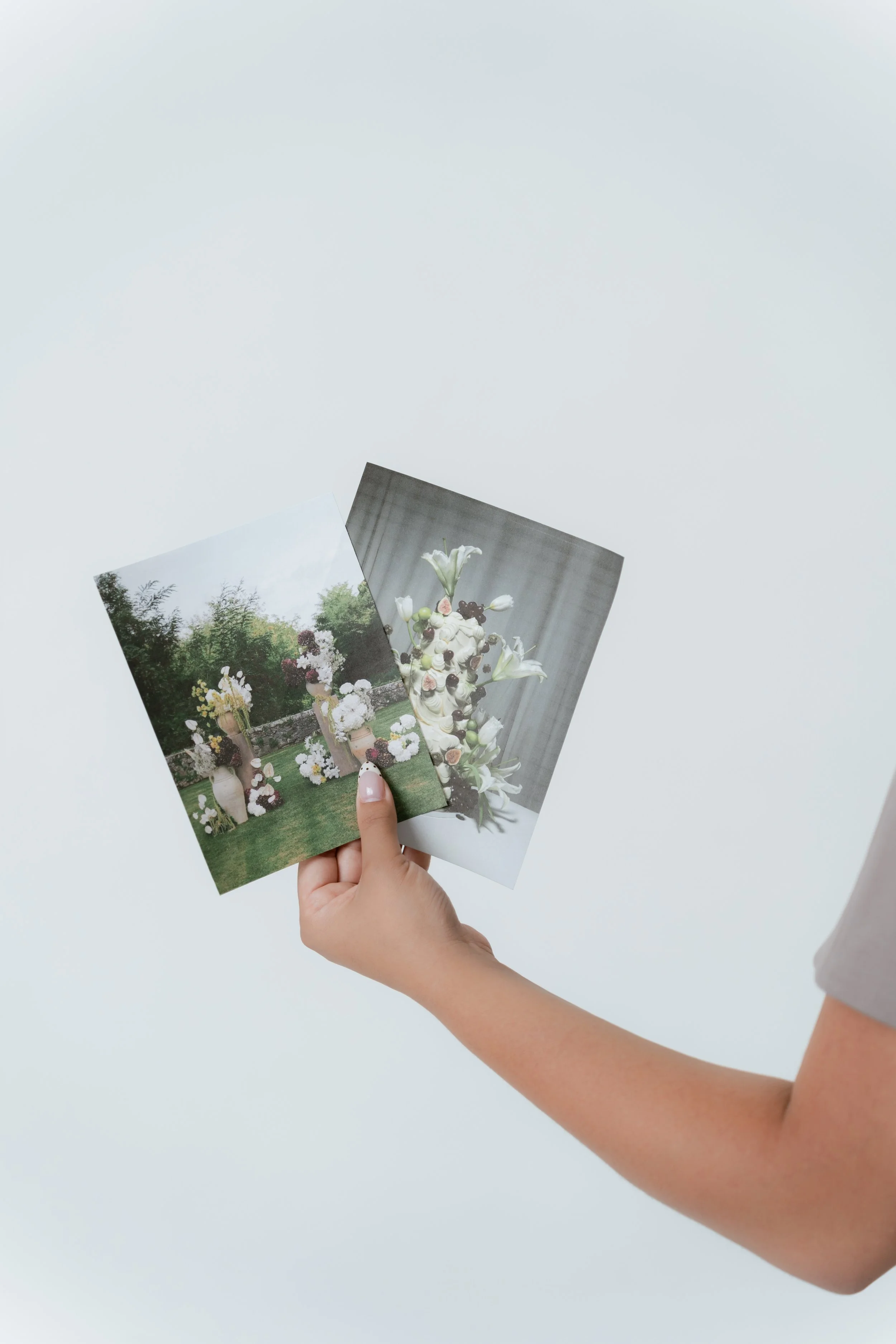 Person holding two photographs of flower arrangements against a plain white background.