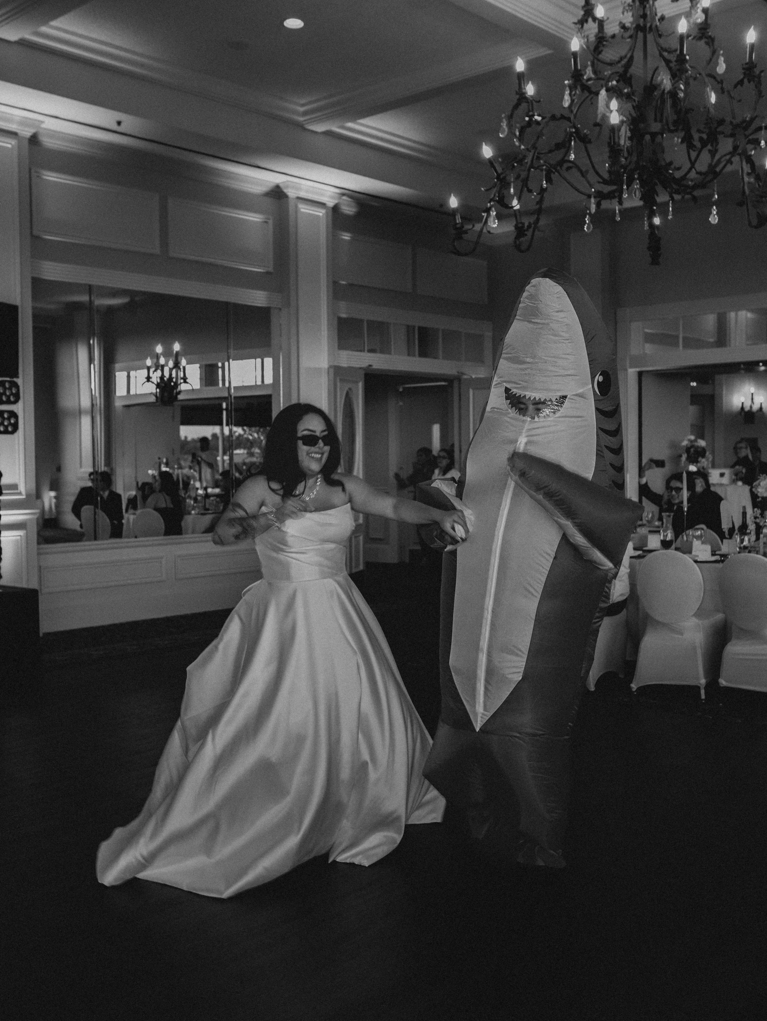 A person in a wedding dress dancing with a person in an inflatable shark costume at a reception hall with chandeliers and large mirrors.