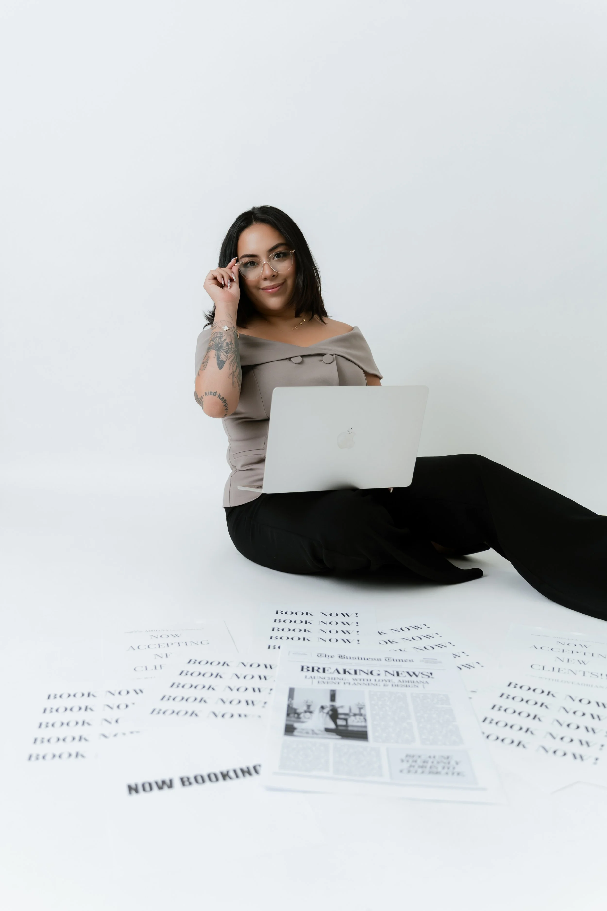 Woman sitting cross-legged on the floor with papers and a laptop, adjusting glasses, against a plain white background.
