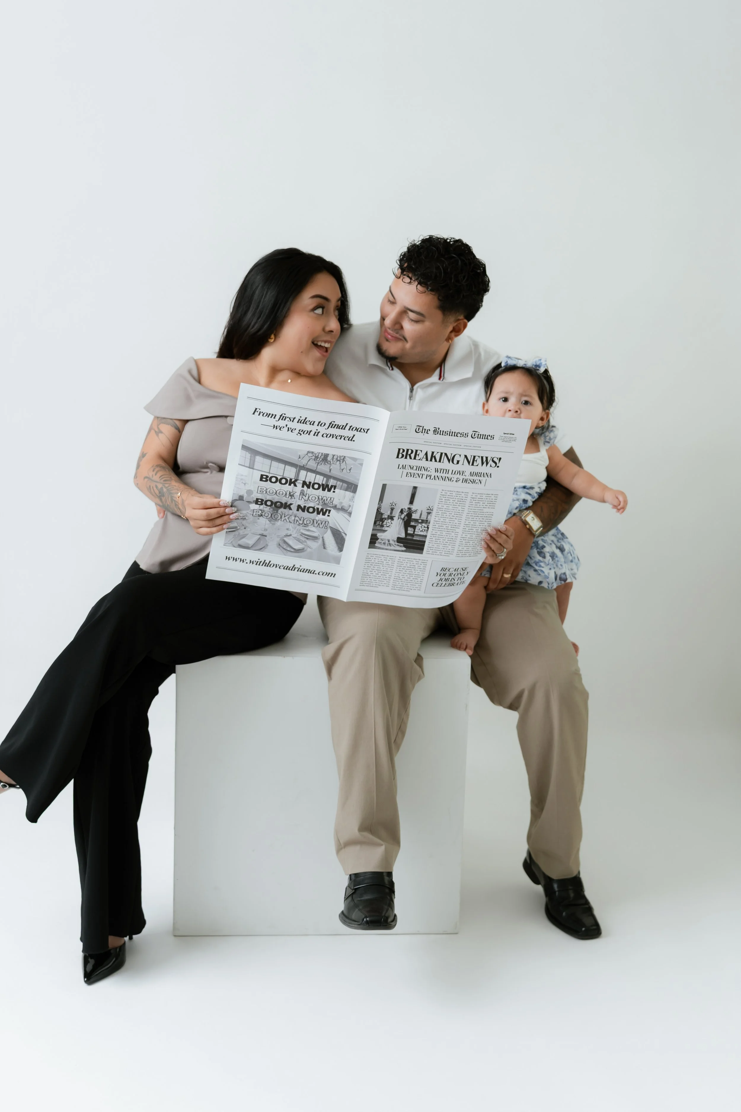 A diverse family of three reading a newspaper together against a plain white background. The woman has tattoos and is smiling, the man is holding the woman and baby, and the baby is looking at the camera.