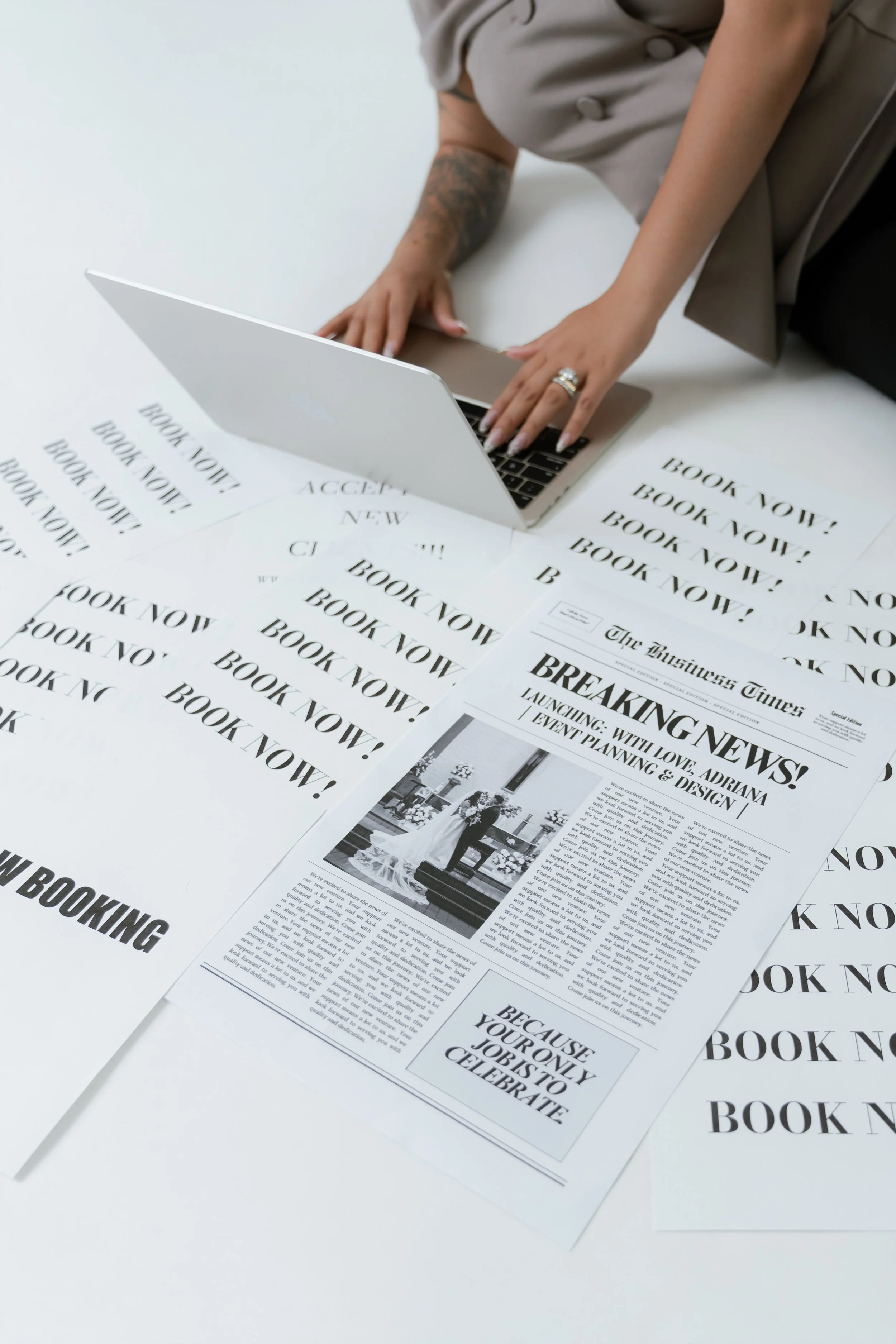 A hand typing on a laptop surrounded by printed newspapers with headlines about booking and breaking news.