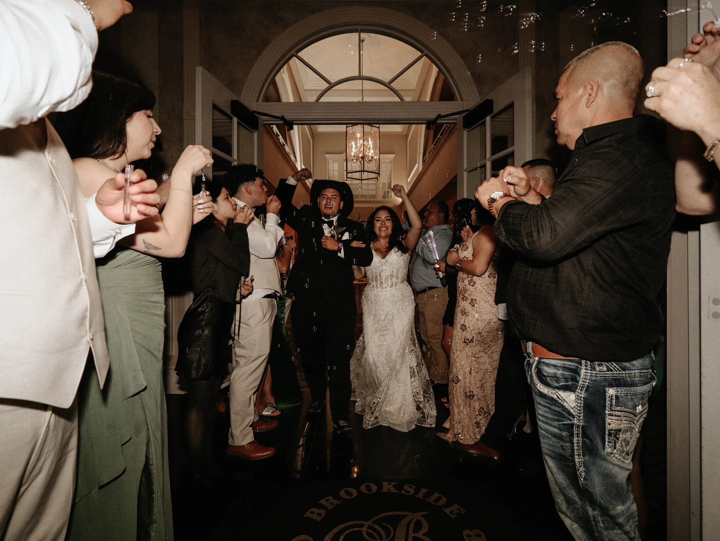 People celebrating at a wedding reception, dancing and cheering inside a grand hall with ornate ceiling details and a chandelier.