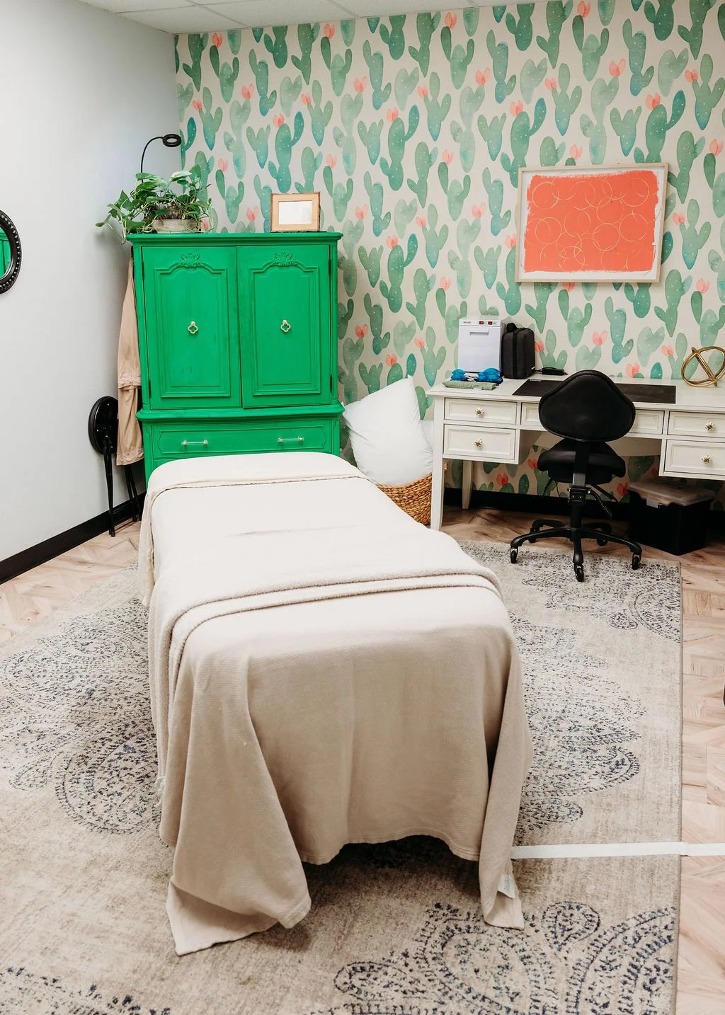 A cozy room with a massage table covered in a beige blanket, a white pillow in a wicker basket, a white desk with a black office chair, a green armoire with plants and a picture on top, and a cactus-patterned wallpaper with pink flowers.