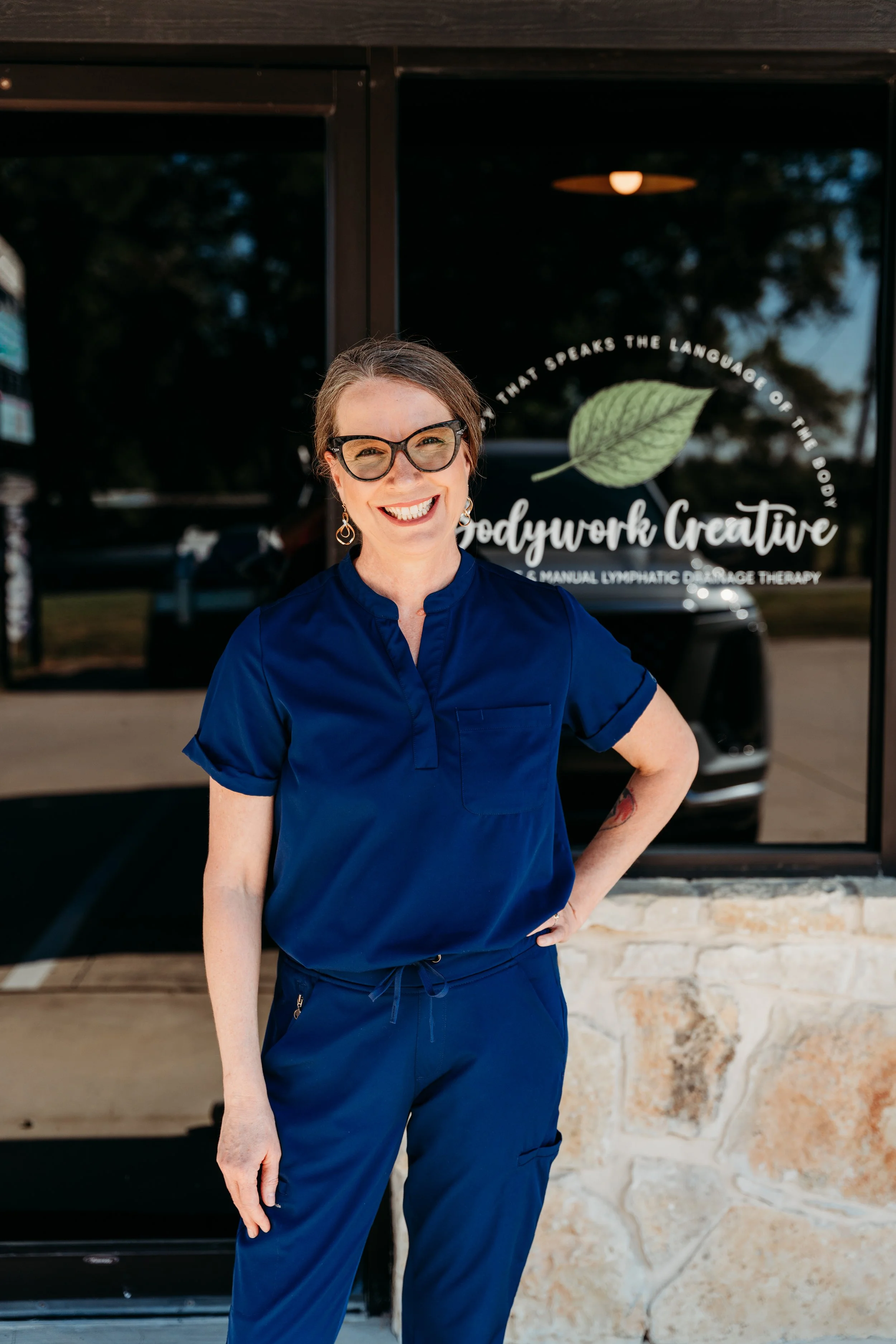 Smiling woman with glasses and earrings standing in front of a glass door with the words 'Bodywork Creative' and a leaf logo, wearing a navy blue uniform.