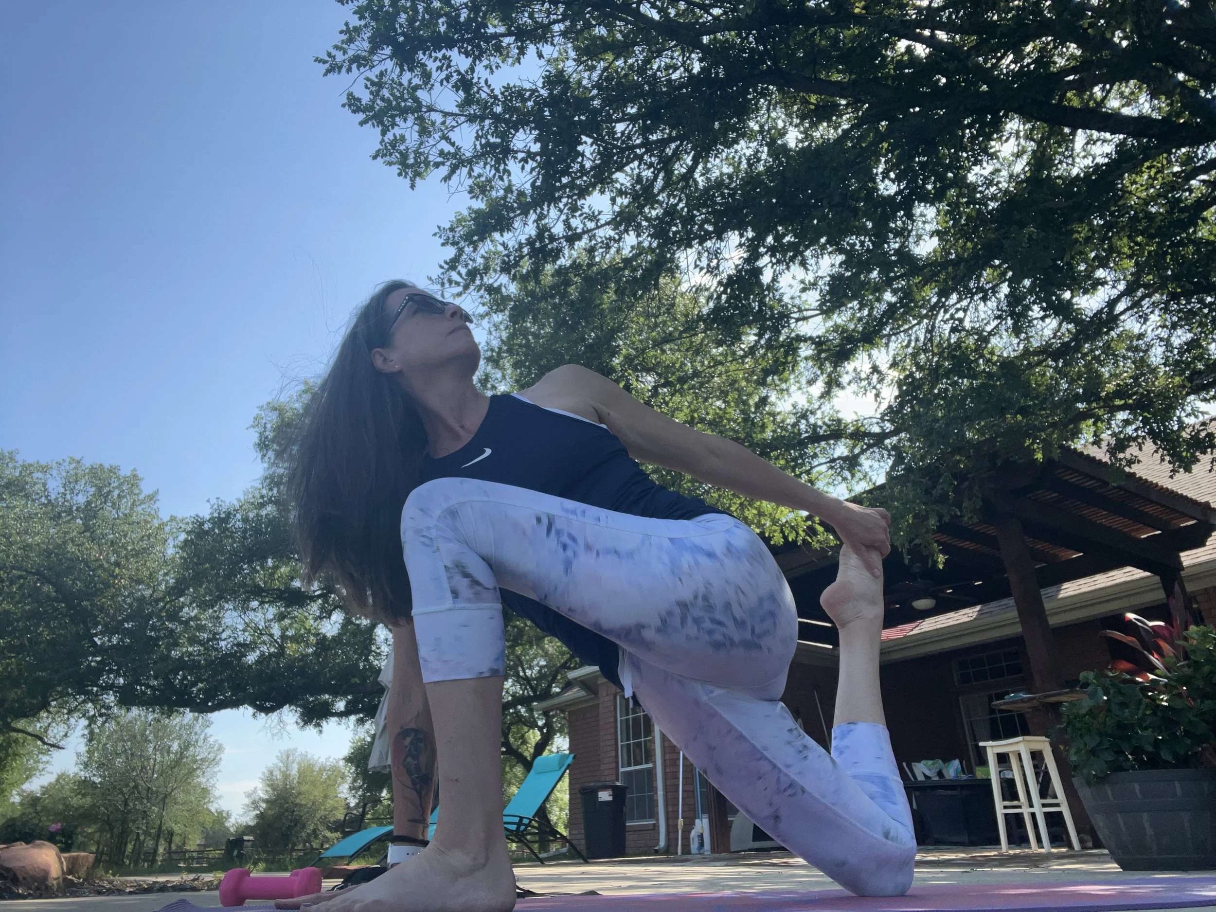 A woman practicing yoga outdoors under a tree, doing a plank pose with her arms and legs extended on a yoga mat.