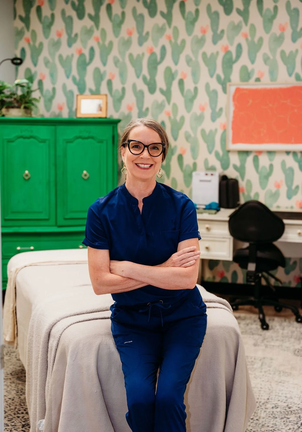 A woman in navy scrubs, wearing glasses, smiling with arms crossed, standing in a colorful room with cactus patterned wallpaper, a green cabinet, and an office chair.