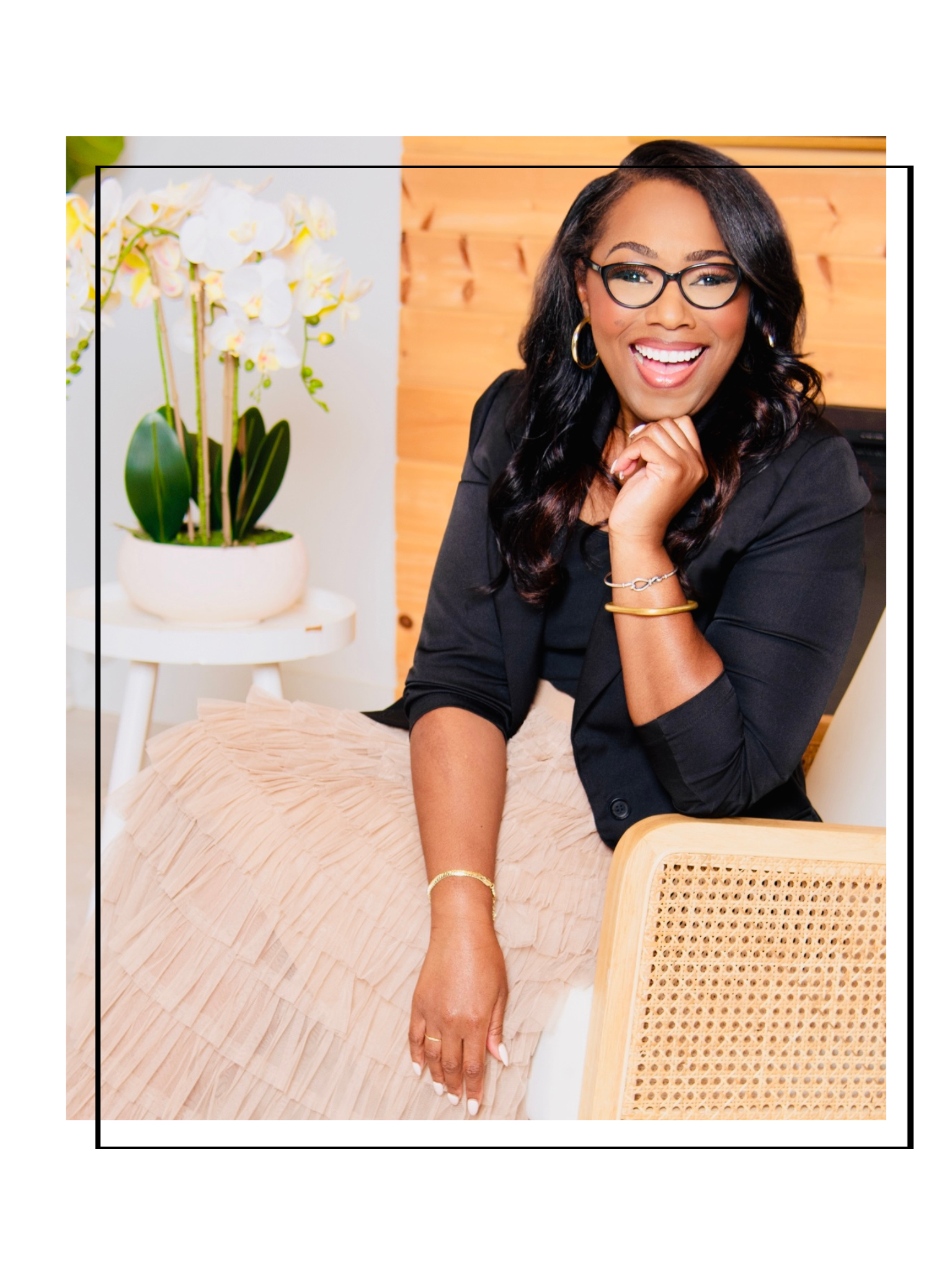 A woman wearing glasses, gold hoop earrings, and a black jacket, smiling with her hand near her face, seated next to a white round table with a potted orchid plant, in a room with wooden wall decor.