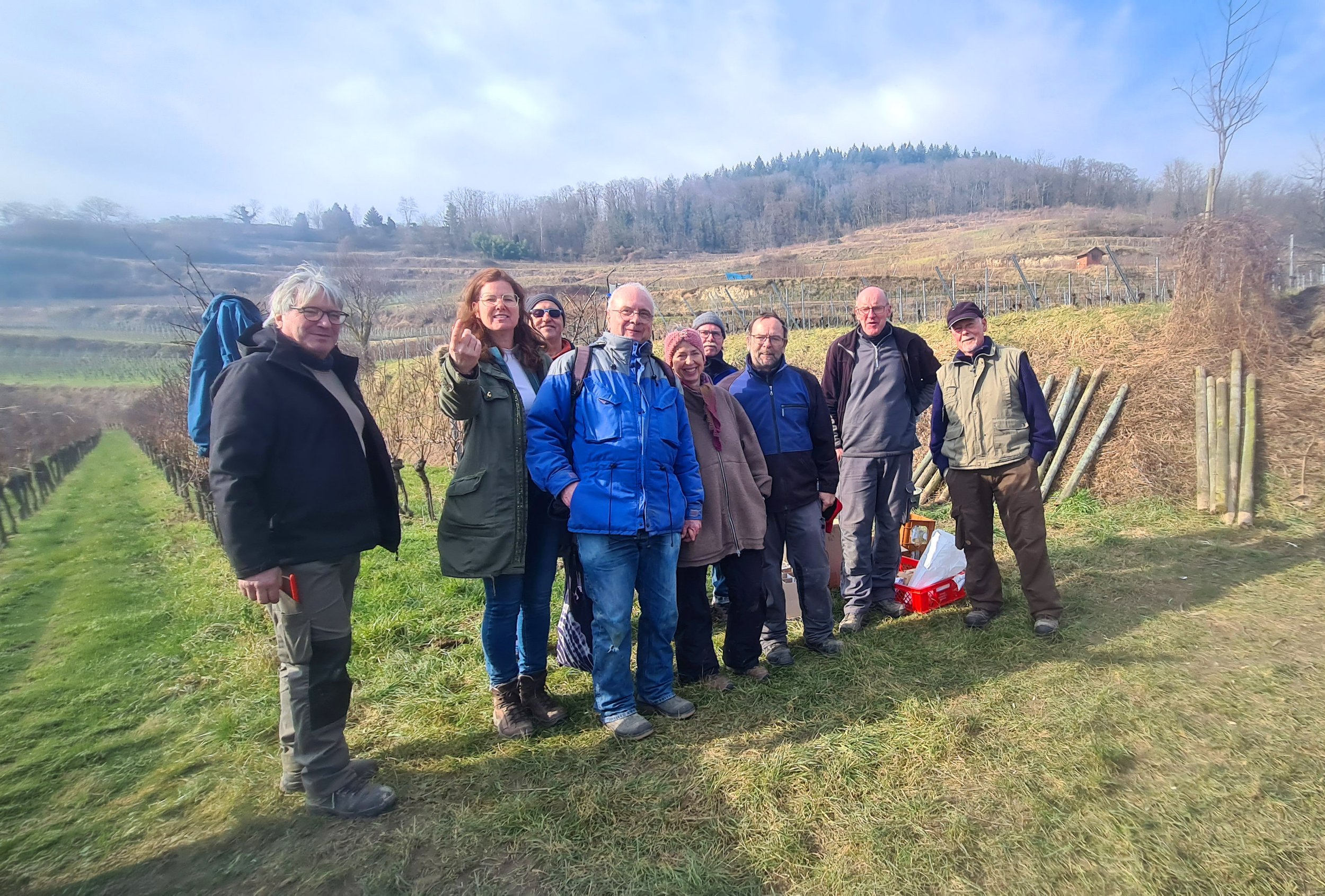 Eine Gruppe von acht Menschen steht in einer Weinberglandschaft, einige tragen Brillen und Jacken, hinter ihnen sind terrassierte Weinberge und Baumgruppen am Hang sichtbar.