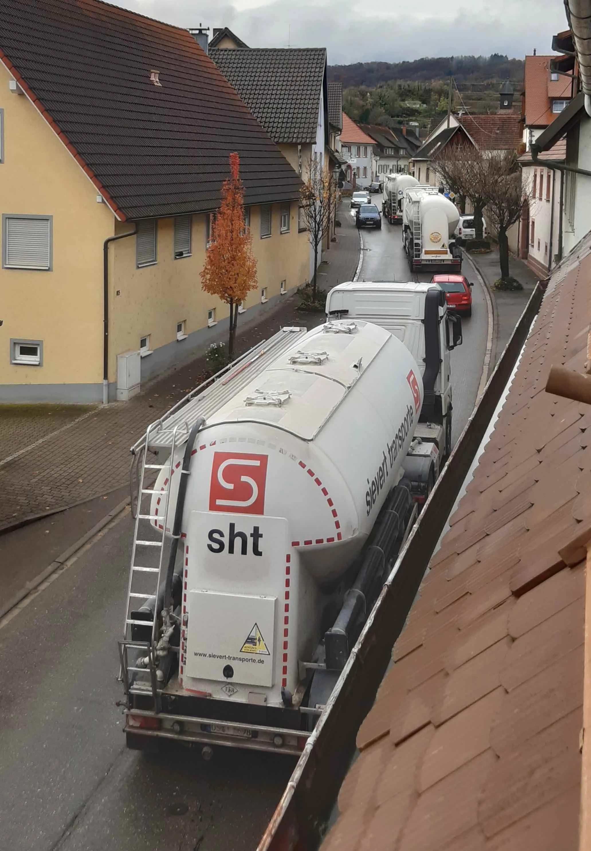 Ein Lastwagen parkt am Straßenrand in einer kleinen Stadt, umgeben von Häusern mit roten und grauen Ziegeldächern. Es ist Herbst, die Bäume haben orange und braune Blätter. Die Straße ist schmal, und der Himmel ist bewölkt.