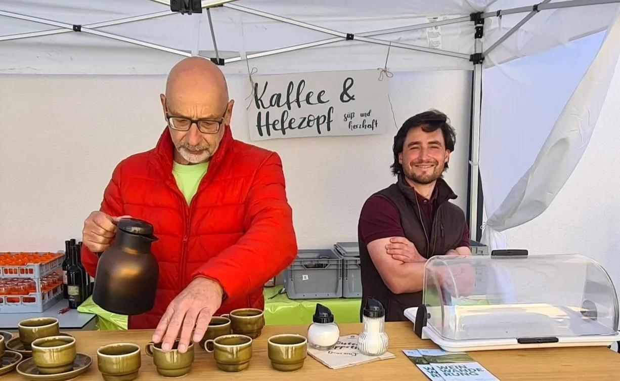 Zwei Männer stehen an einem Verkaufsstand. Der ältere Mann in rotem Jacke gießt Kaffee in Tassen, während der jüngere Mann lächelnd daneben steht. Der Stand hat eine Tafel mit der Aufschrift 'Kaffe & Hefezoop' und weitere Zeichen.