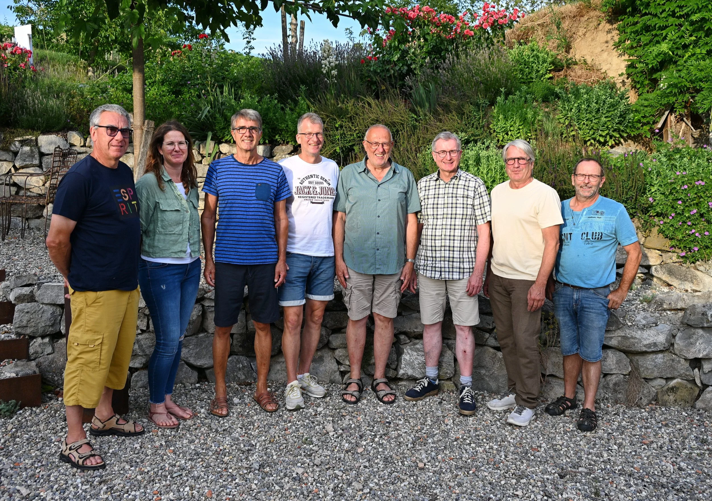 Gruppenfoto von neun Personen, die im Freien vor einer Steinmauer und einem üppigen Blumen- und Gartengürtel stehen, bei Sonnenschein.
