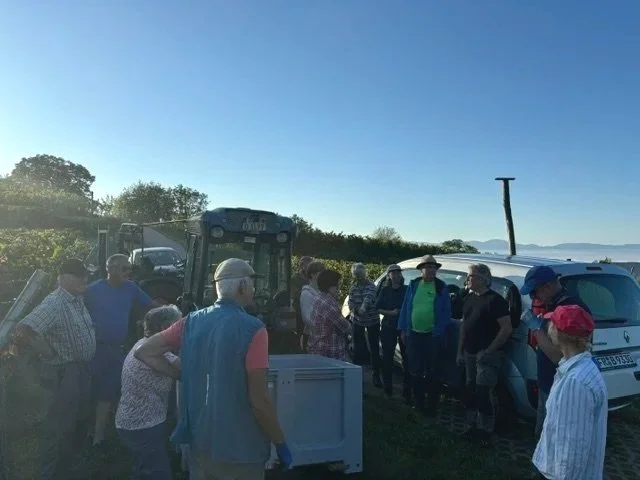 Mehrere Menschen versammeln sich draußen an einem sonnigen Tag mit einem Traktor, einem Auto und einem Wasserstandsholz in einer ländlichen Umgebung.