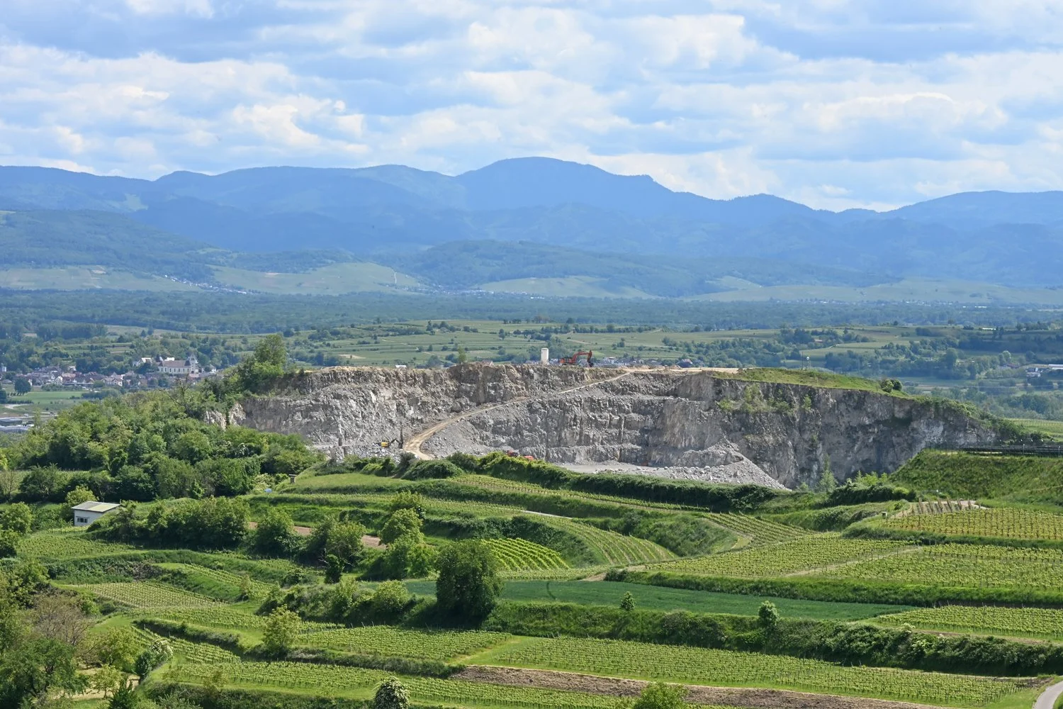 Landschaft mit Weinbergen im Vordergrund, einer Steinbruchansicht in der Mitte und Bergen im Hintergrund, unter einem bewölkten Himmel.