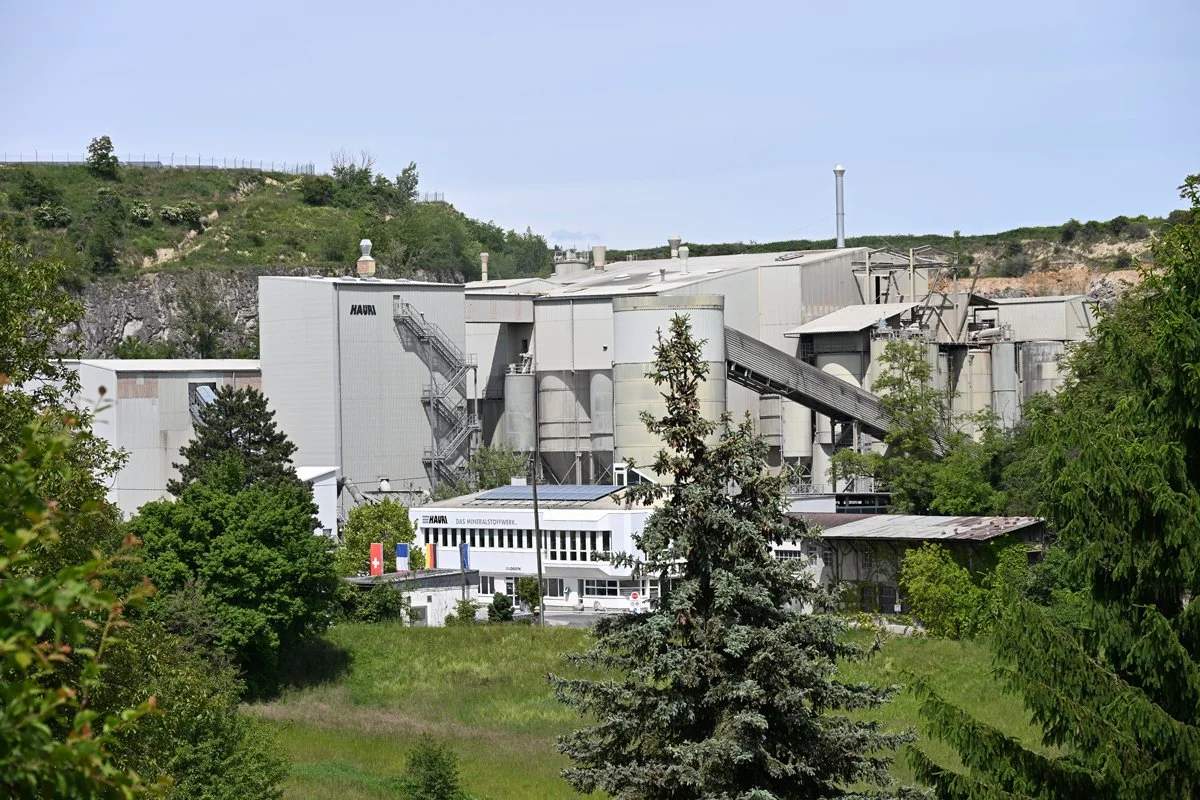 Ein großes Industriegebäude, wahrscheinlich eine Mineralstofffabrik, umgeben von Bäumen und grünen Hügeln im Hintergrund.