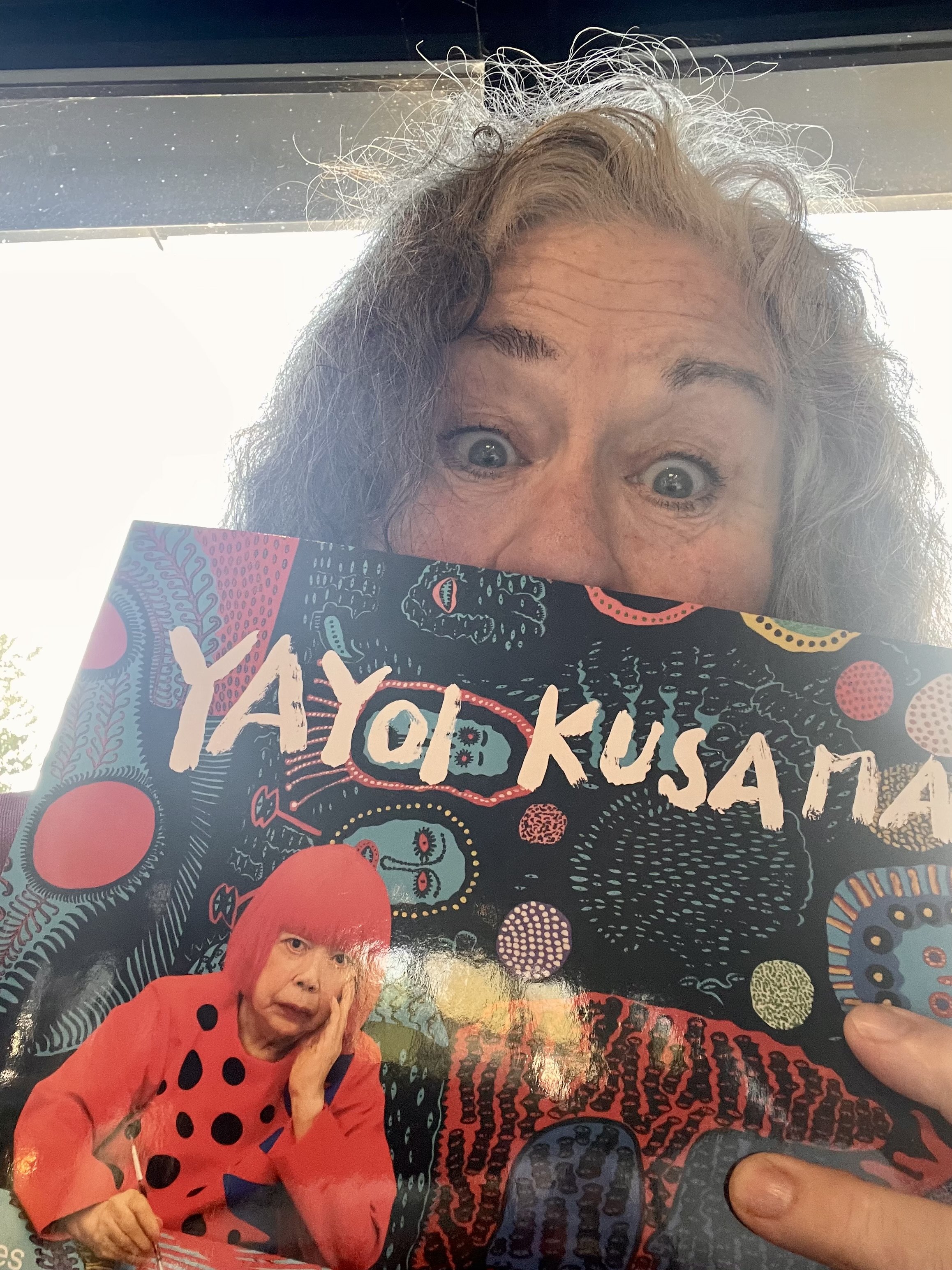 Close-up of a woman with curly gray hair holding a colorful book titled "AYOKA" with graphic illustrations; she is looking over the top of the book with wide eyes and a playful expression.
