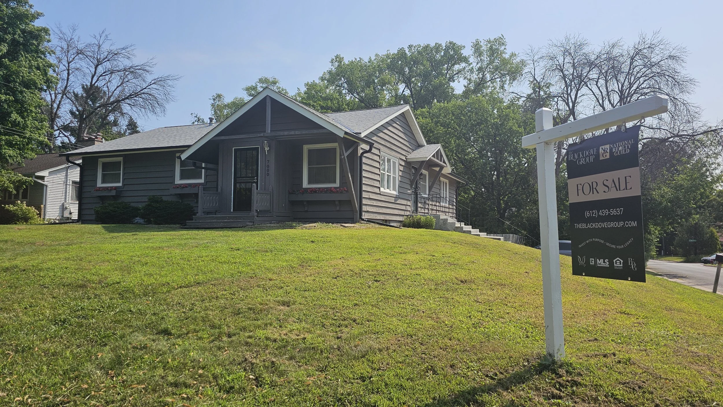A gray house with a front porch and stairs, surrounded by a green lawn and trees, with a for-sale sign in the yard.