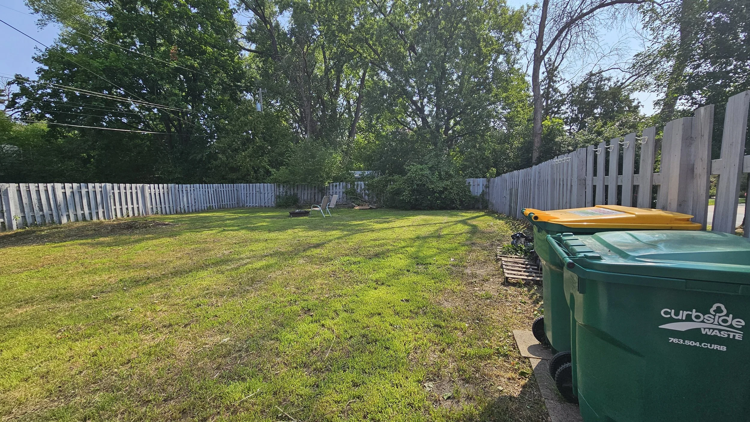 Residential backyard with green grass, a white wooden fence, and large trees. Two colorful trash bins (green and yellow) are in the foreground, with a black trash bin labeled 'curbside waste' nearby. Two patio chairs and a small fire pit are visible near the fence.