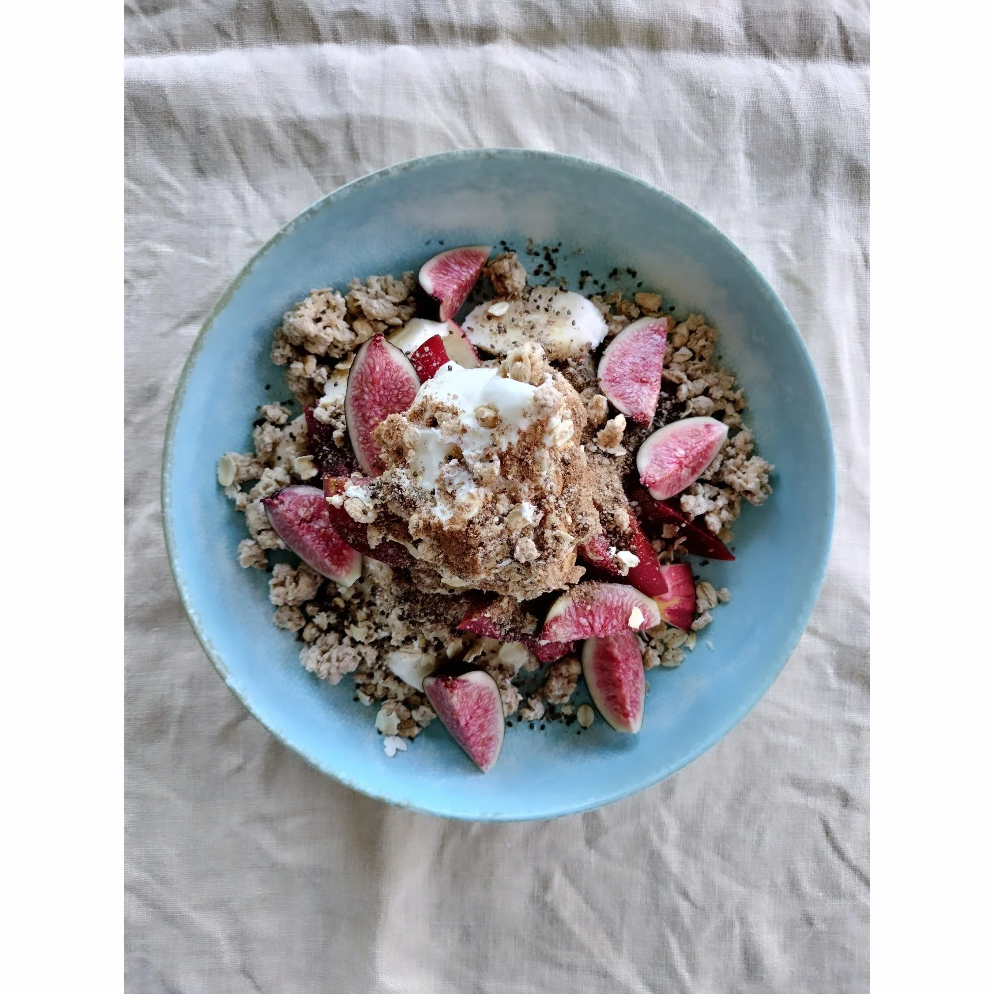 A light blue bowl containing granola, sliced peaches, a dollop of yogurt, and black chia seeds, placed on a white fabric surface.