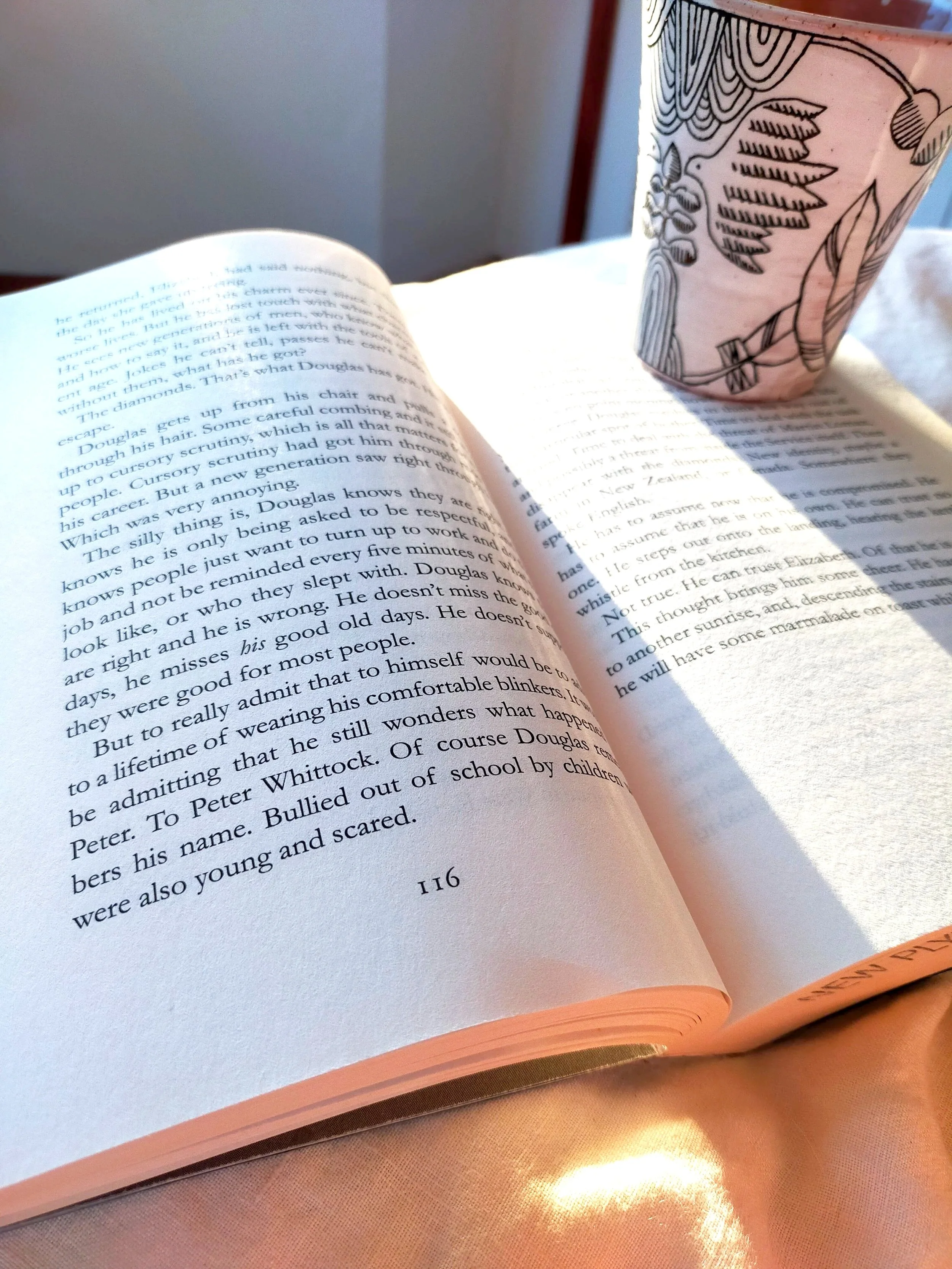 An open book resting on a pink surface with a pink mug decorated with black line drawings of leaves and a bird positioned nearby. The pages of the book contain printed text.