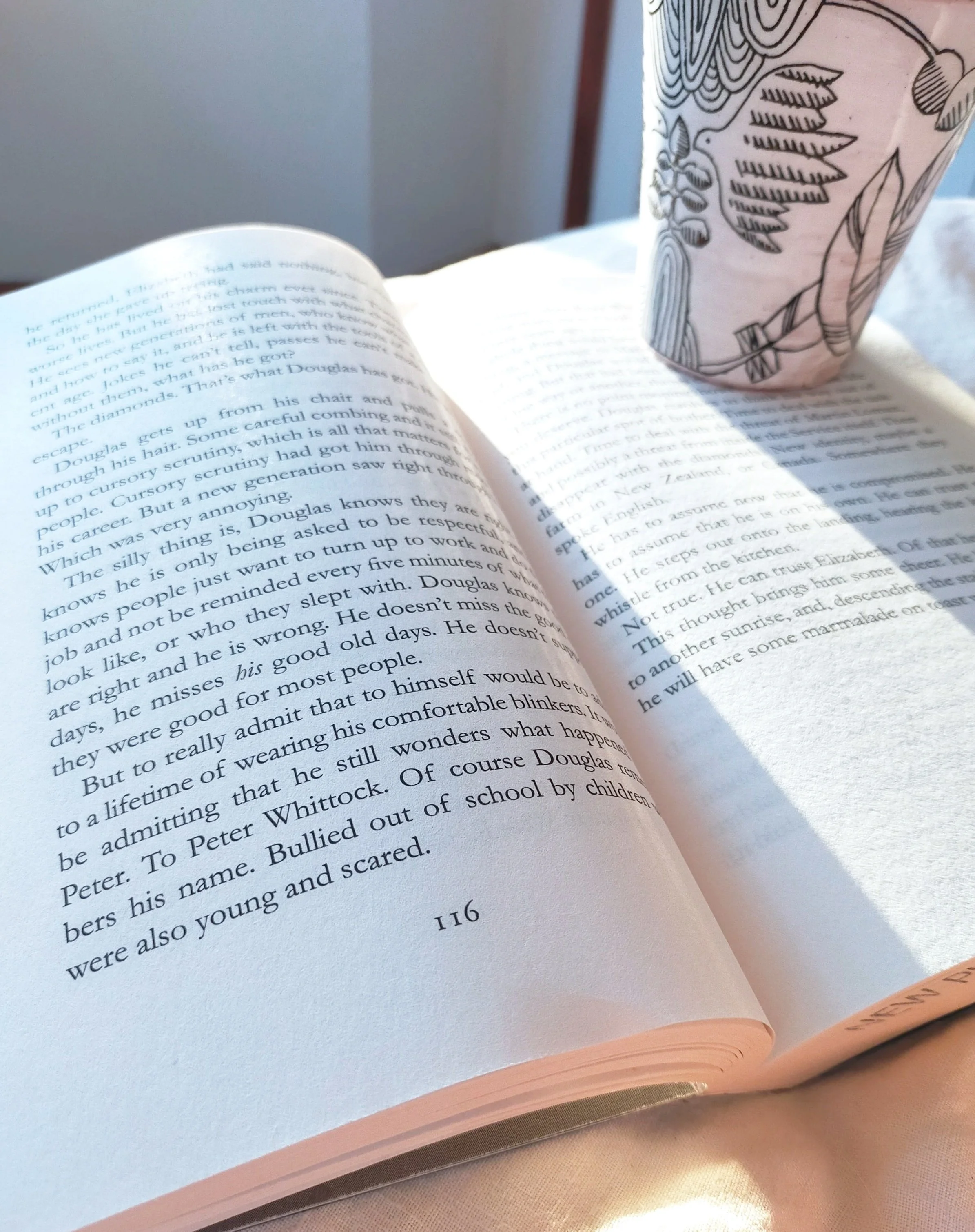 Open book with printed text and a paper cup with a black outline floral pattern on a table.