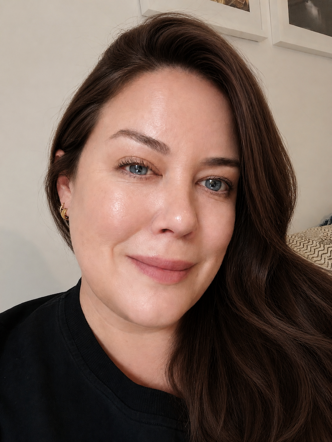 Close-up of a woman with long, wavy brown hair, blue eyes, light skin, wearing small gold earrings and a black top, smiling softly in an indoor setting with framed pictures on the wall in the background.